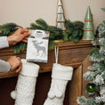 Load image into Gallery viewer, A person places a Fresh Scents Woodland Sachet 3pk above a wooden mantel decorated with pine branches, ceramic trees, and white knit stockings. This flameless fragrance adds a cozy touch near the frosted Christmas tree.
