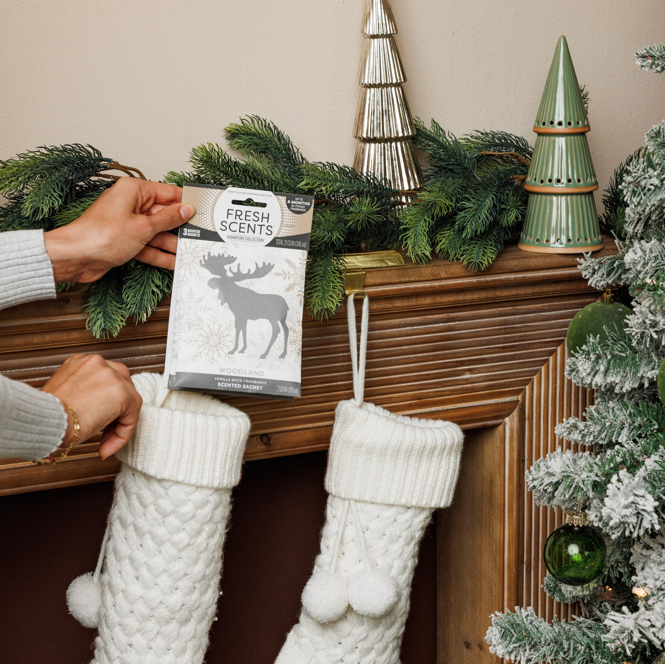 A person places a Fresh Scents Woodland Sachet 3pk above a wooden mantel decorated with pine branches, ceramic trees, and white knit stockings. This flameless fragrance adds a cozy touch near the frosted Christmas tree.