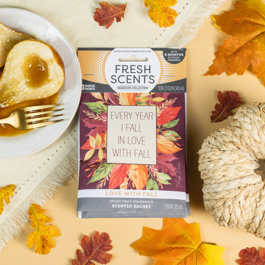 The Fresh Scents Love with Fall - Sachet 3pk, adorned with "Every year I fall in love with fall," sits on a yellow background among autumn leaves, a white pumpkin, and caramel pears—a charming flameless fragrance for your space.