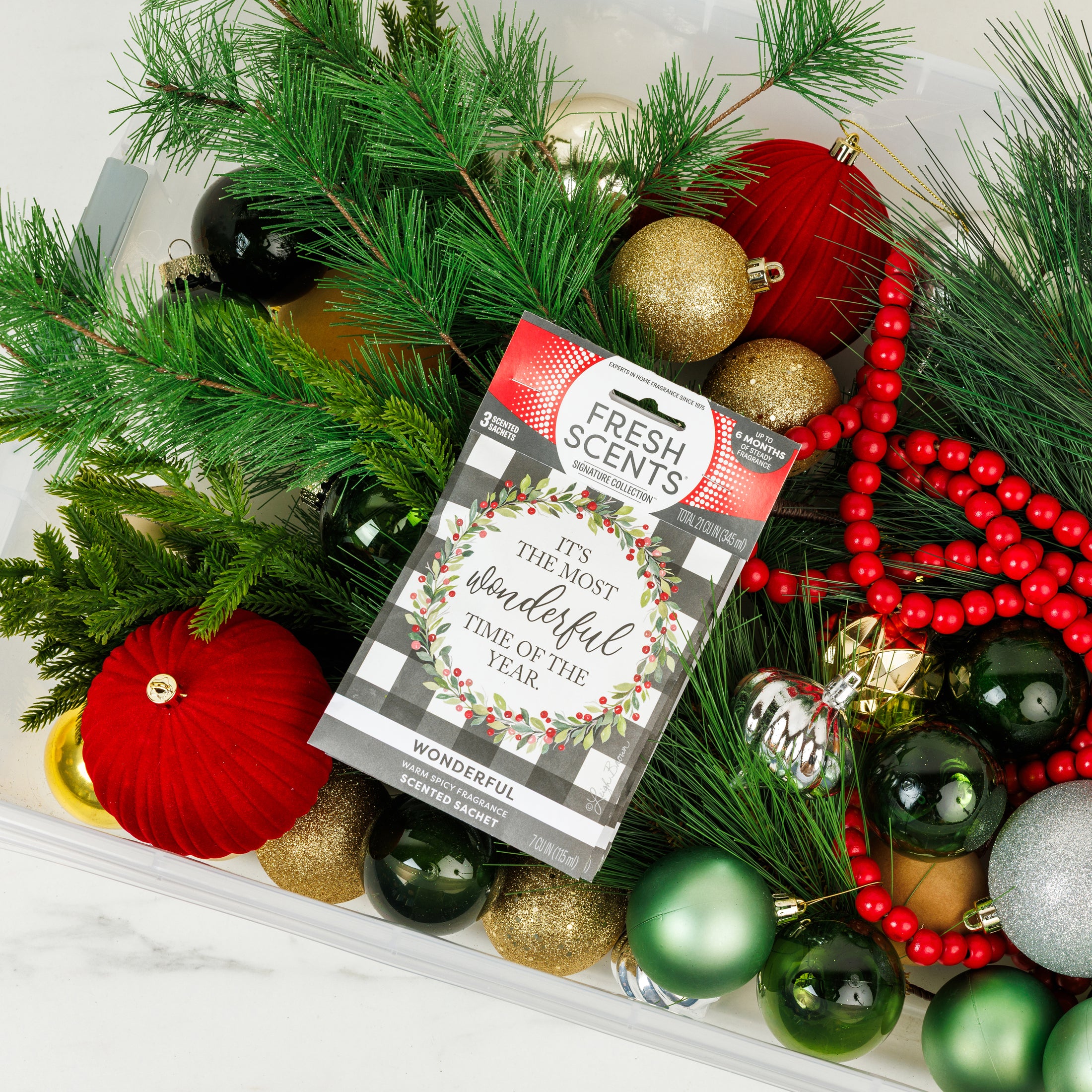 A plastic bin filled with Christmas decorations—green pine branches, red and gold ornaments, red beads, and a Fresh Scents "Wonderful - Sachet 3pk" air freshener labeled “It’s the most wonderful time of the year.”.