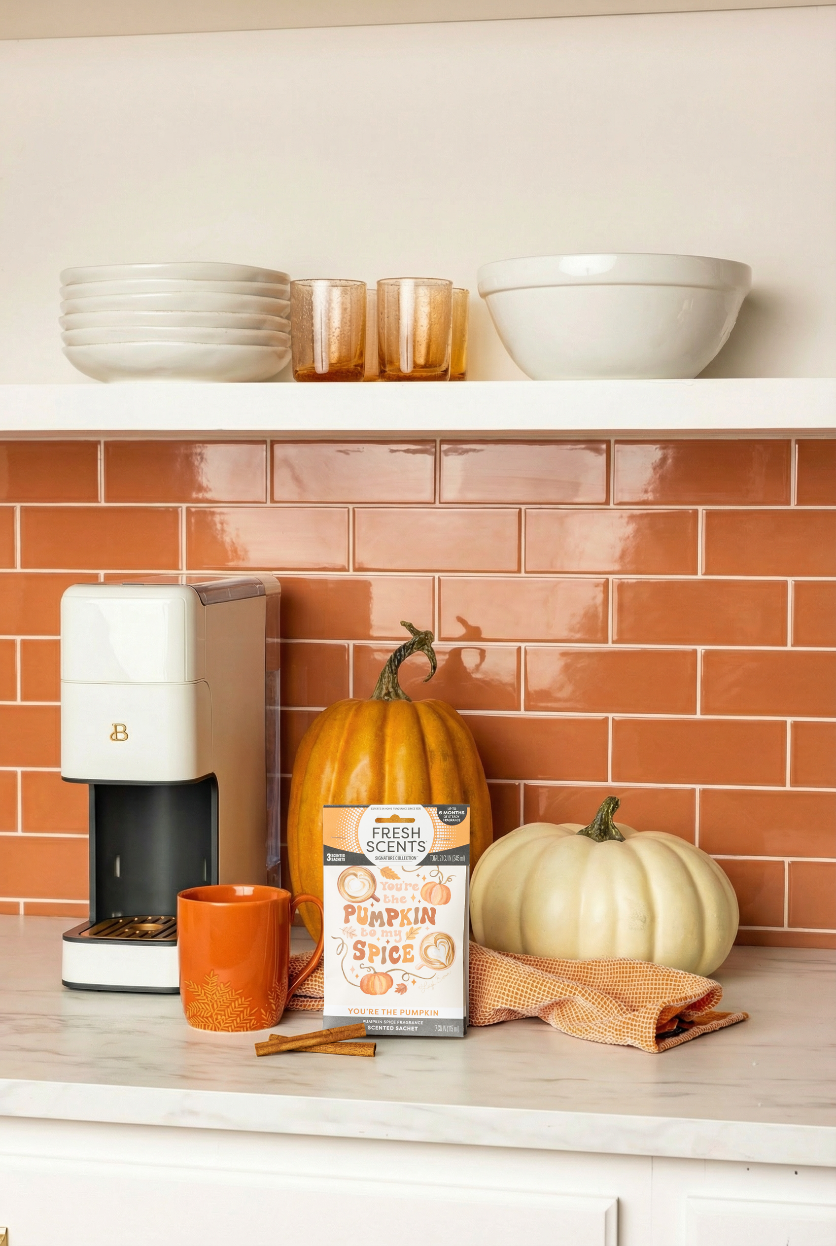 A kitchen counter displays a coffee maker, an orange mug, a pack of Fresh Scents “You’re the Pumpkin - Sachet 3pk,” two pumpkins (orange and white), and a folded orange towel. White dishes and glasses sit on shelves above an orange tile backsplash.