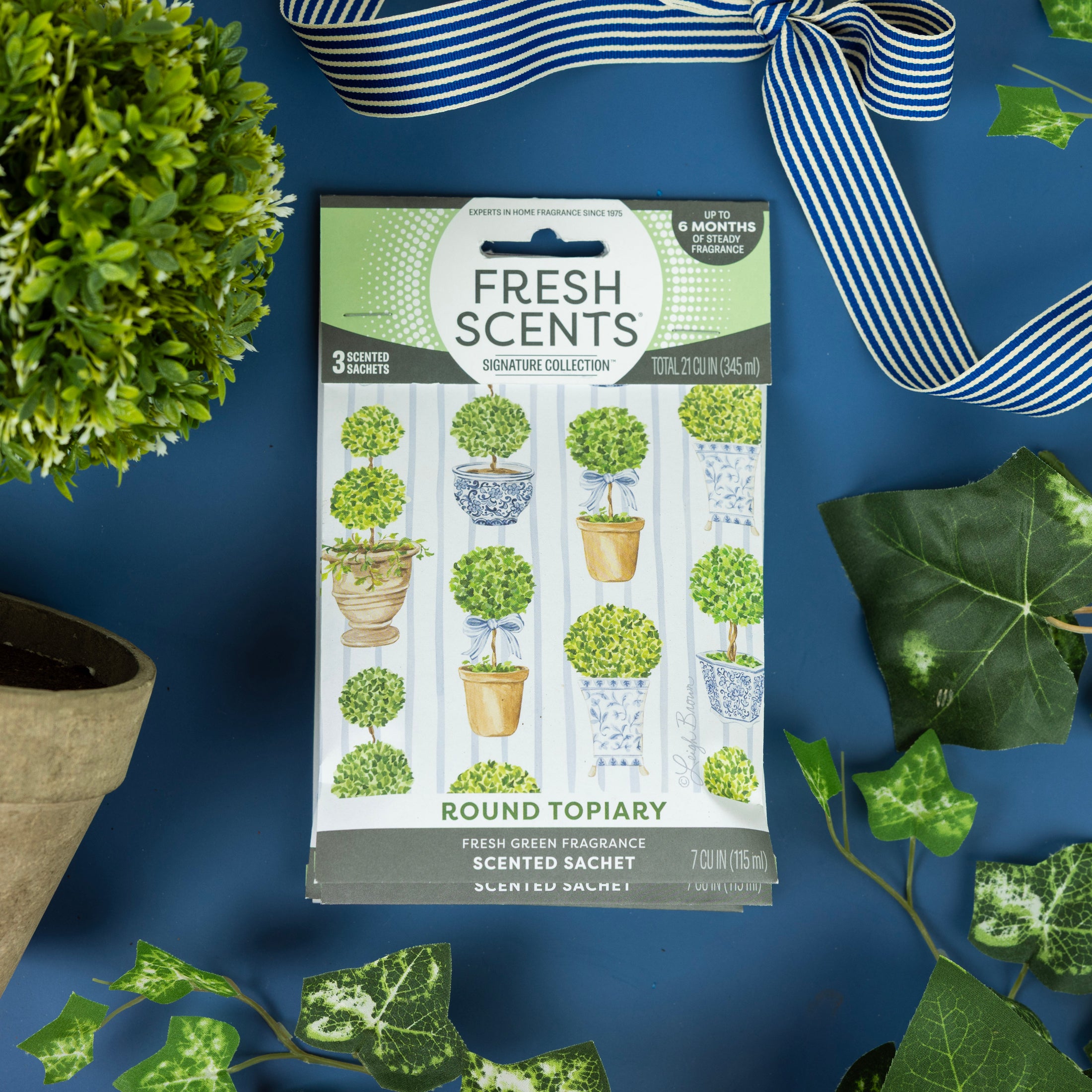 A package of Fresh Scents Round Topiary - Sachet 3pk rests on a blue surface, surrounded by potted plants, ivy leaves, and a blue-and-white striped ribbon. The packaging displays illustrations of topiary plants.