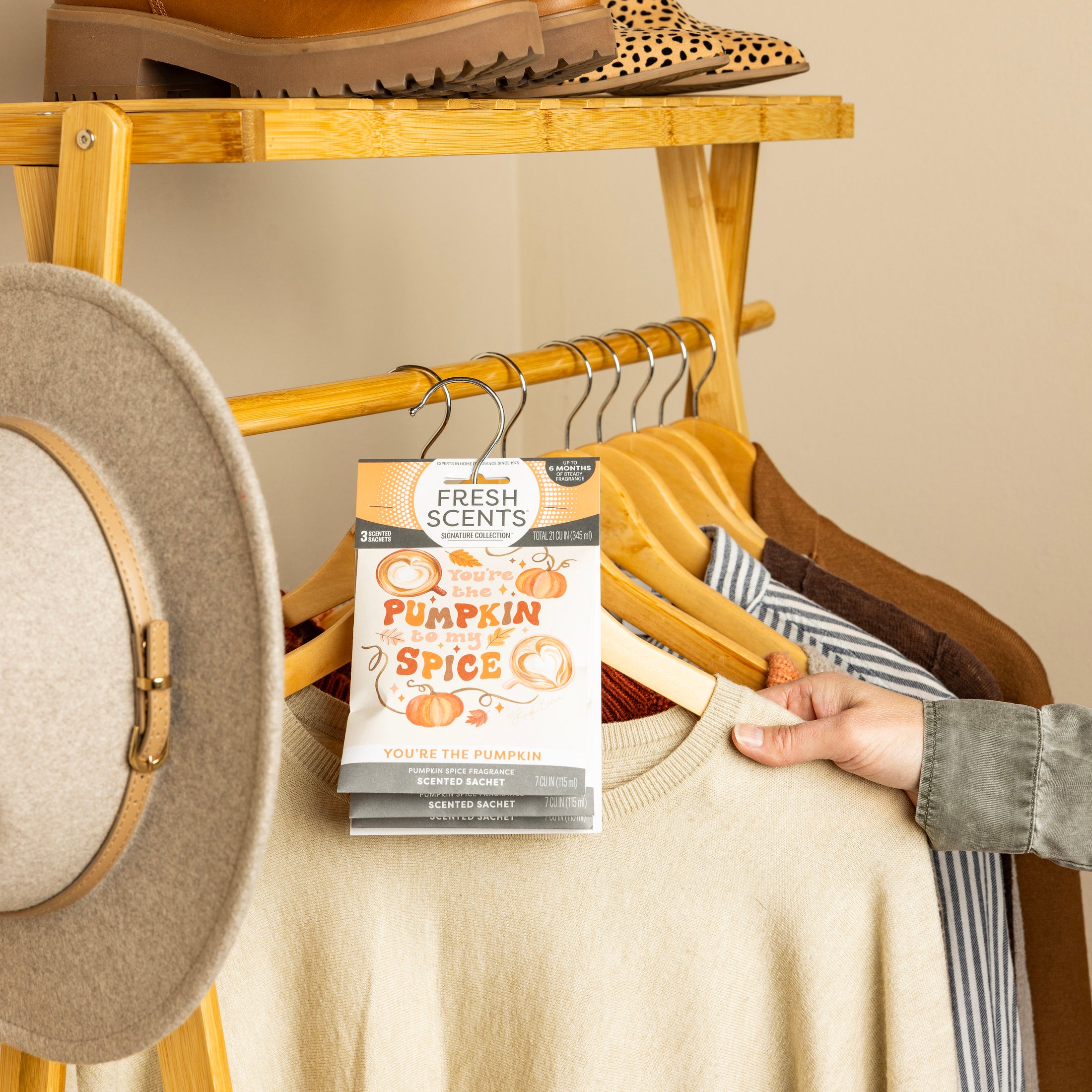 A hand hangs a Fresh Scents You're the Pumpkin - Sachet 3pk on a wooden rack with sweaters, a tan hat, and brown shoes—a charming flameless air freshener for long-lasting fragrance.