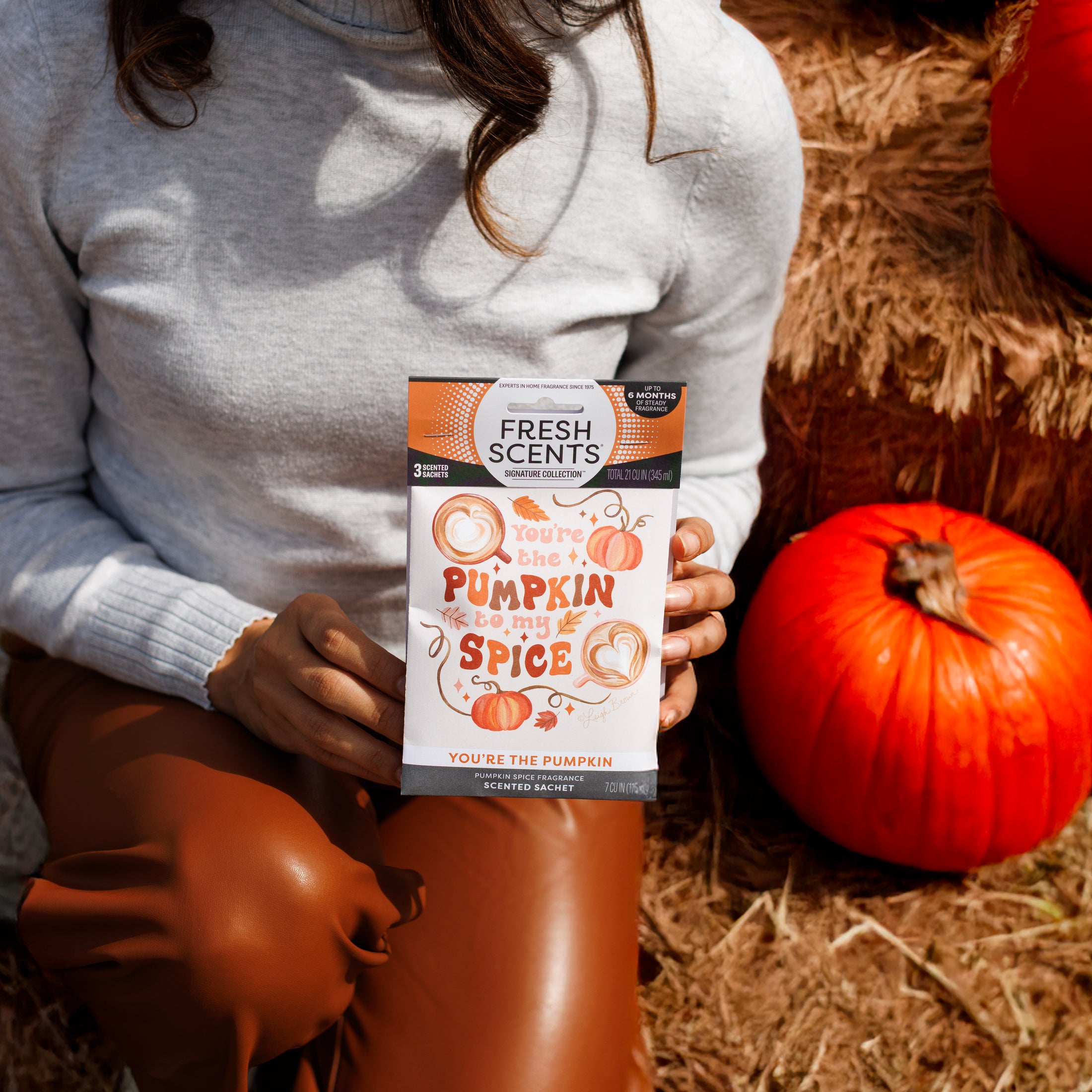 A person in a light gray sweater and brown pants sits on hay with orange pumpkins, holding a Fresh Scents You're the Pumpkin - Sachet 3pk.