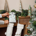 Load image into Gallery viewer, A person puts a Fresh Scents Balsam Fir - Sachet 3pk into a white knitted stocking hanging on a festive mantel with green garland and trees, beside a snowy Christmas tree decorated with green ornaments.
