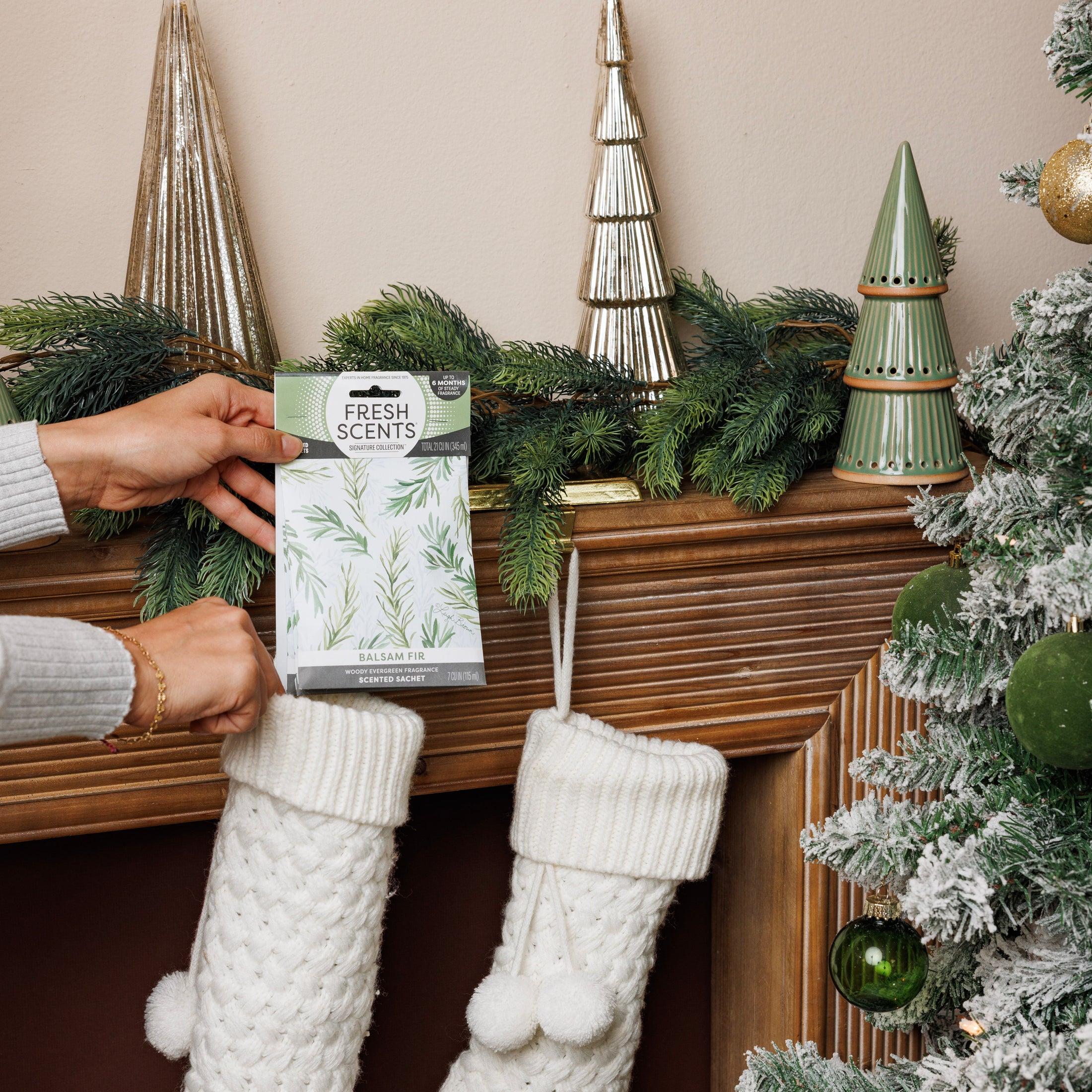 A person puts a Fresh Scents Balsam Fir - Sachet 3pk into a white knitted stocking hanging on a festive mantel with green garland and trees, beside a snowy Christmas tree decorated with green ornaments.