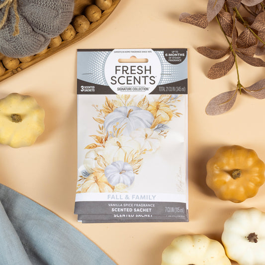 The Fresh Scents Fall & Family - Sachet 3pk, a flameless air freshener with long-lasting fragrance, sits on a tan surface with small white and yellow pumpkins, a blue cloth, knit fabric, and autumn leaves.