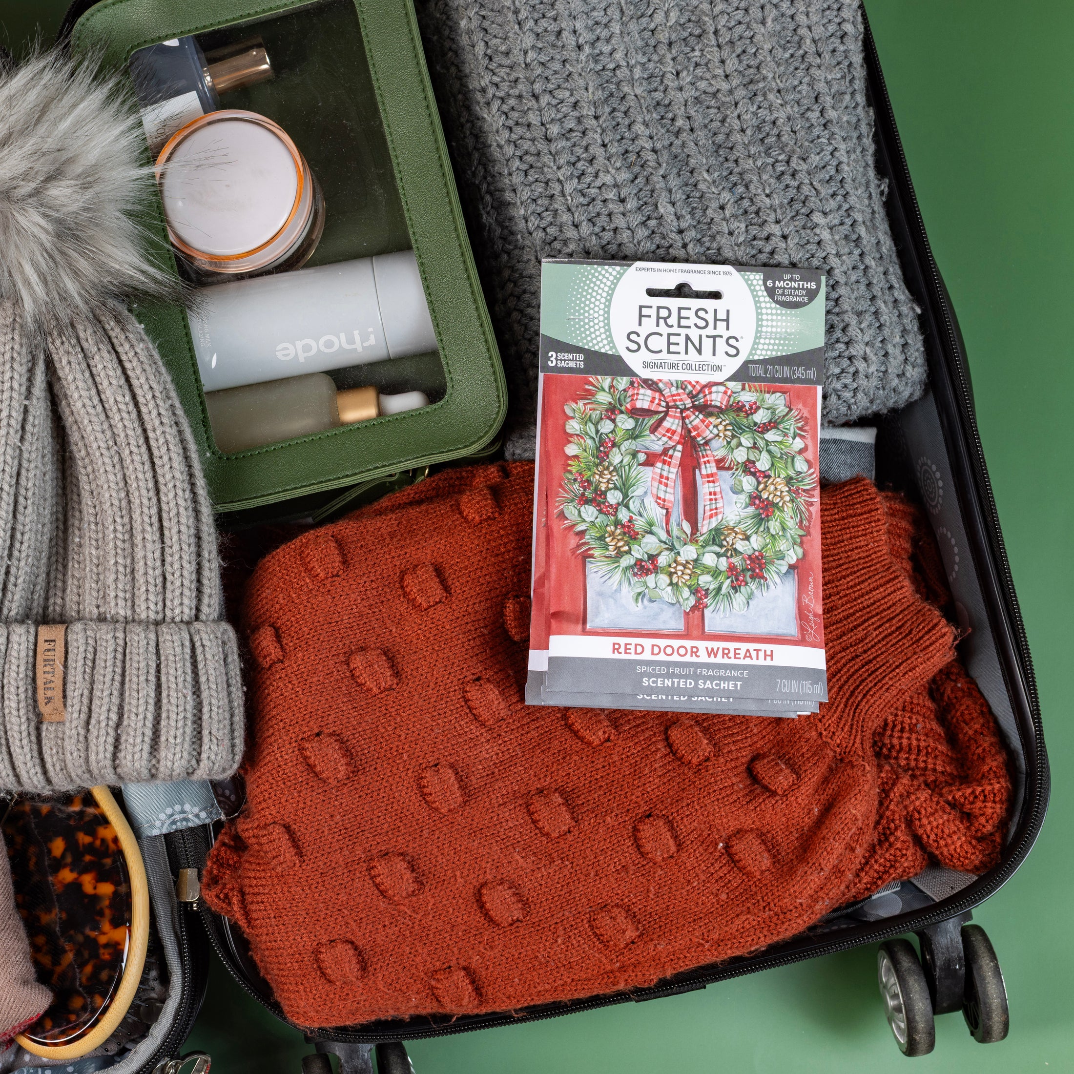 An open suitcase contains a gray knit sweater, an orange patterned sweater, a gray beanie, tortoiseshell sunglasses, a green toiletry case, and a Fresh Scents Red Door Wreath - Sachet 3pk for flameless fragrance.