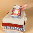 Load image into Gallery viewer, A hand places a Fresh Scents Love with Fall - Sachet 3pk on folded sweaters, adding flameless fragrance and a long-lasting air freshener touch against a beige background.
