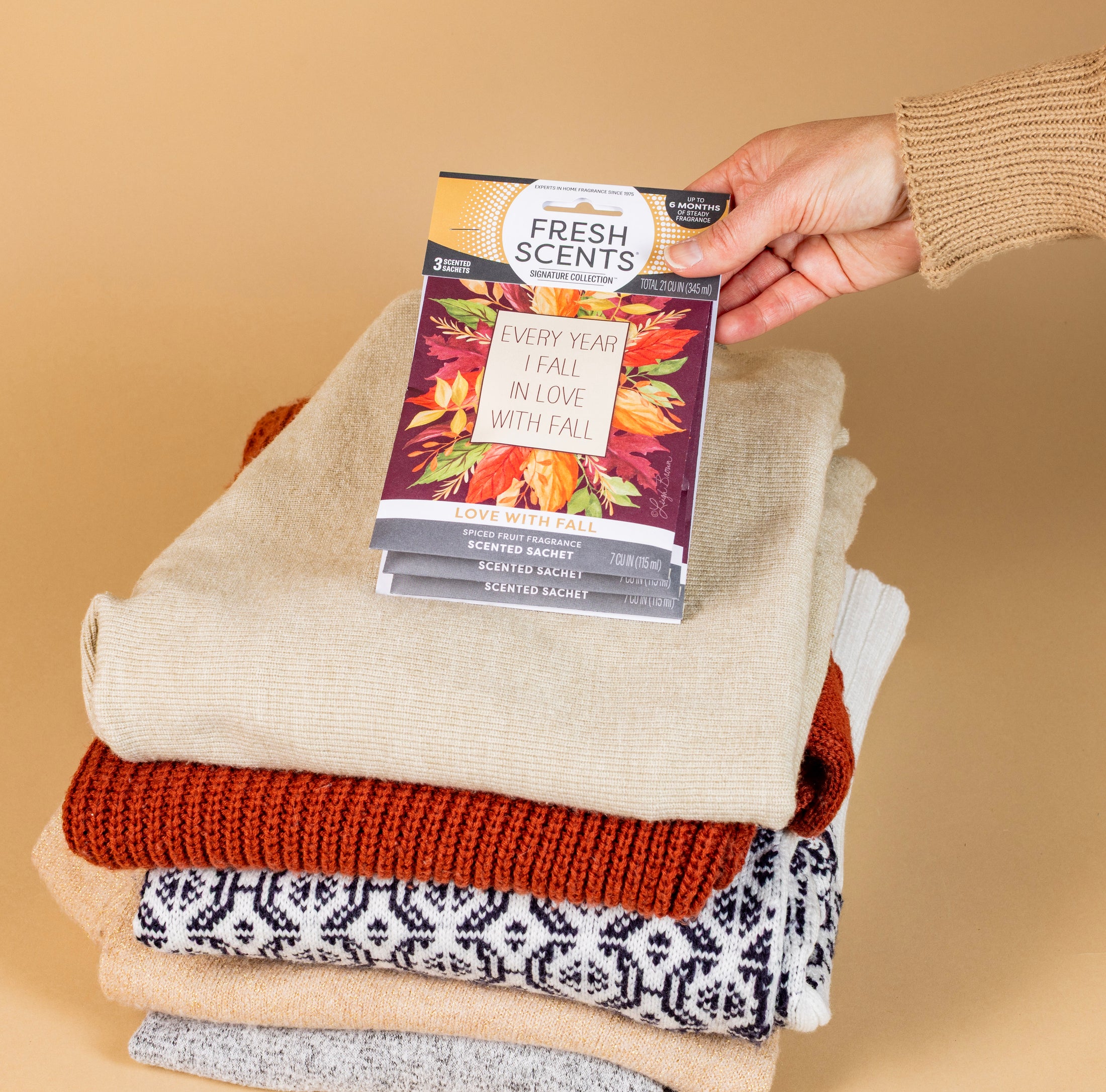A hand places a Fresh Scents Love with Fall - Sachet 3pk on folded sweaters, adding flameless fragrance and a long-lasting air freshener touch against a beige background.