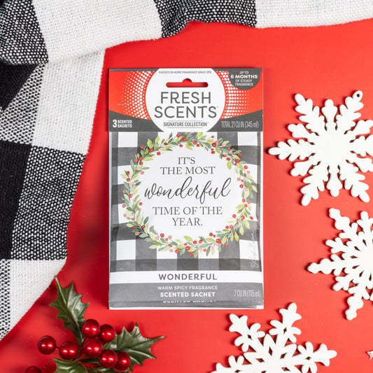 A Fresh Scents Wonderful - Sachet 3pk from the Signature Collection, featuring a wreath and "It's the most wonderful time of the year" text, sits on a red surface with snowflakes, holly, and a black-and-white cloth for lasting fragrance.