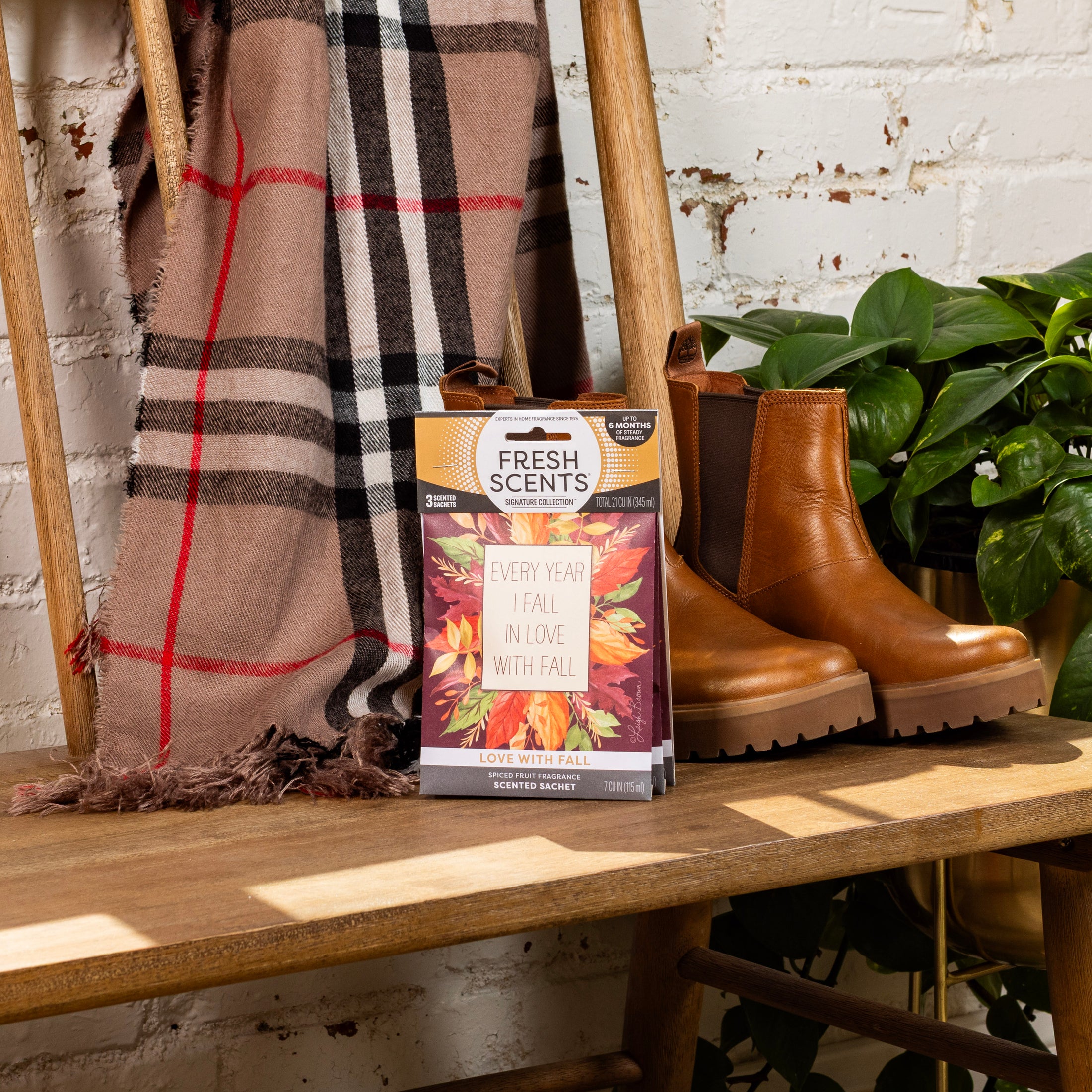 A bench with brown boots, a plaid blanket, a green plant, and a Fresh Scents Love with Fall - Sachet 3pk box decorated with autumn leaves and the phrase "Every year I fall in love with fall.