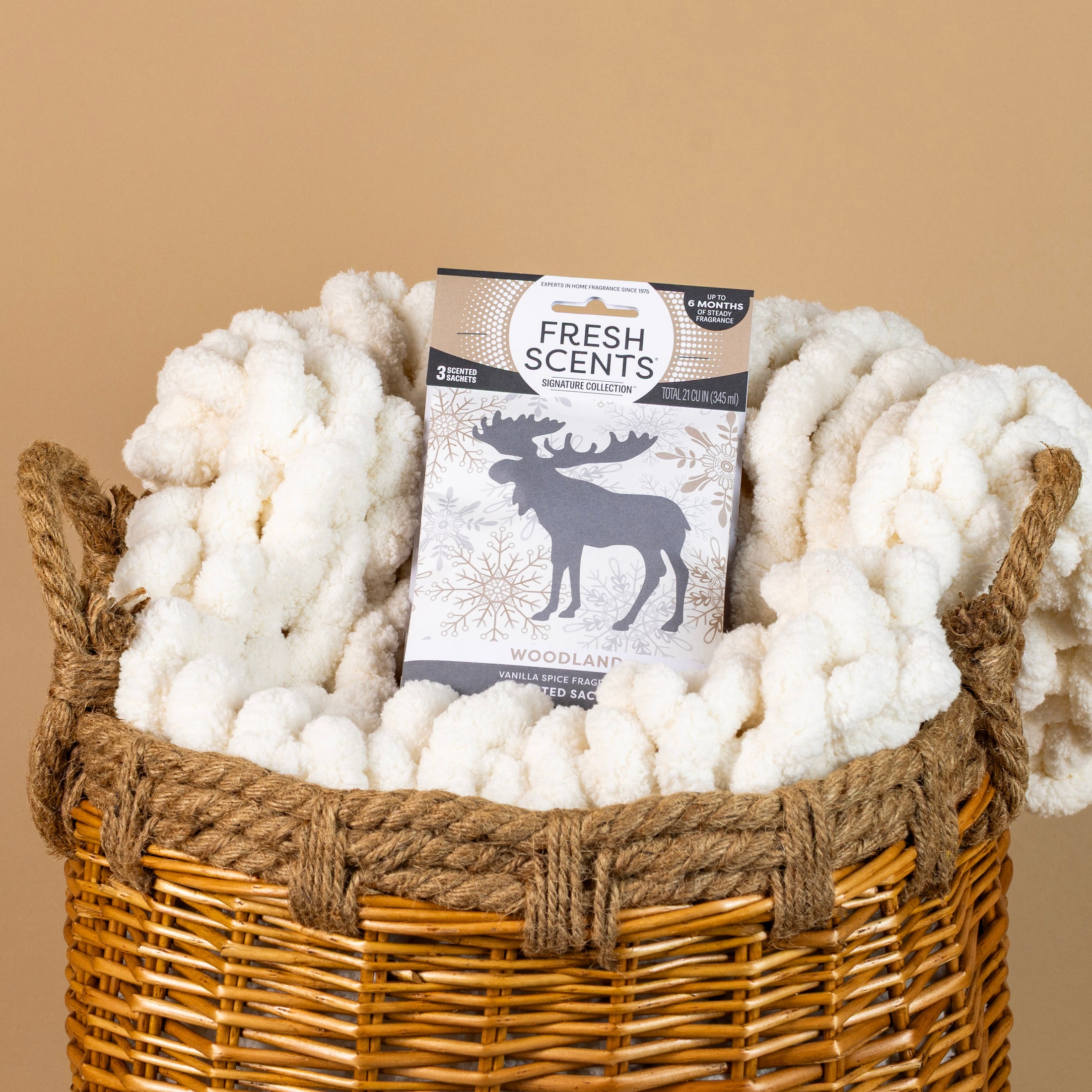 A wicker basket holds a fluffy white blanket and a Fresh Scents Woodland Sachet 3pk, all set against a plain beige background.