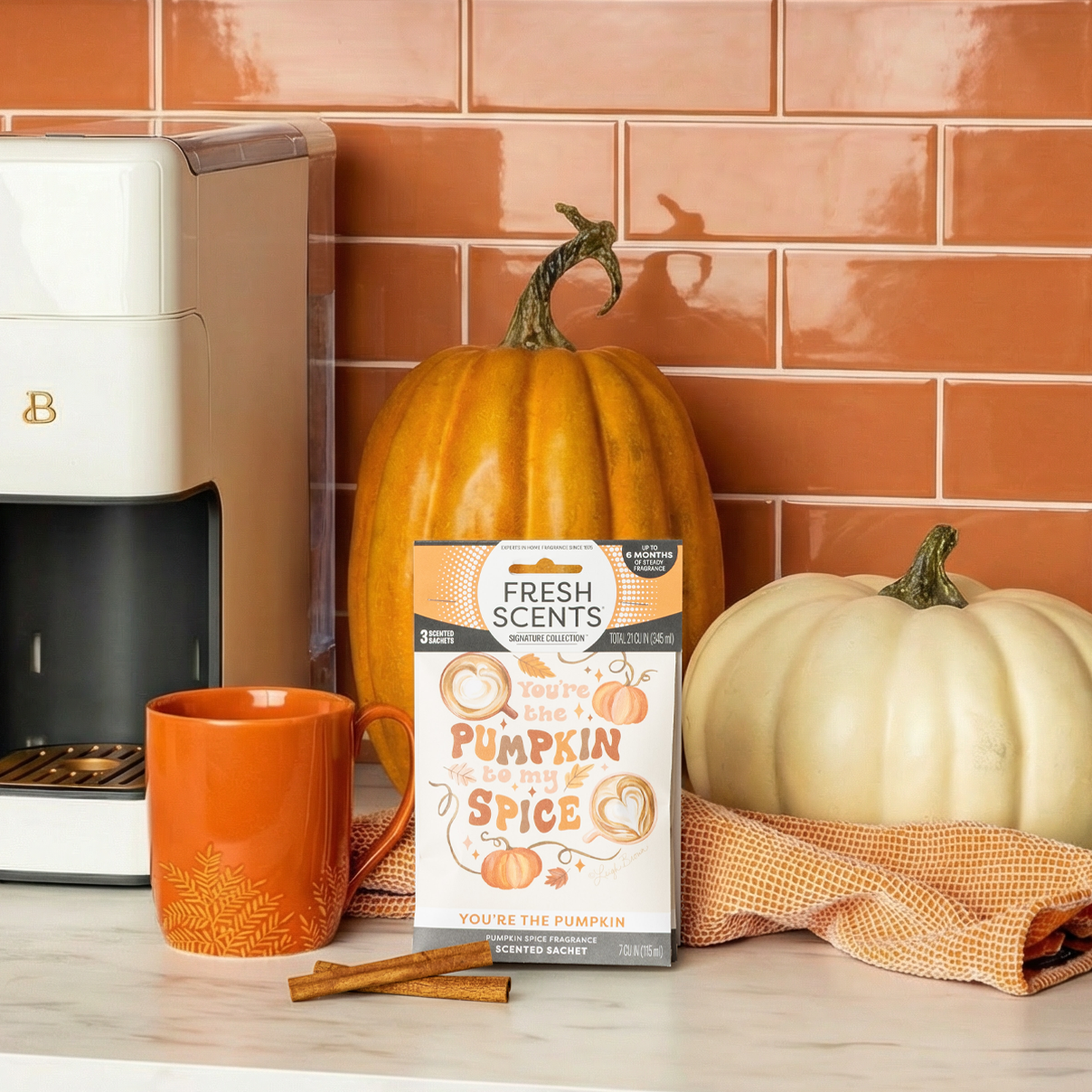 An orange mug, two pumpkins, Fresh Scents "You're the Pumpkin" sachets 3pk with long-lasting fragrance, and cinnamon sticks are arranged on a kitchen counter in front of an orange tiled wall and a white coffee maker.