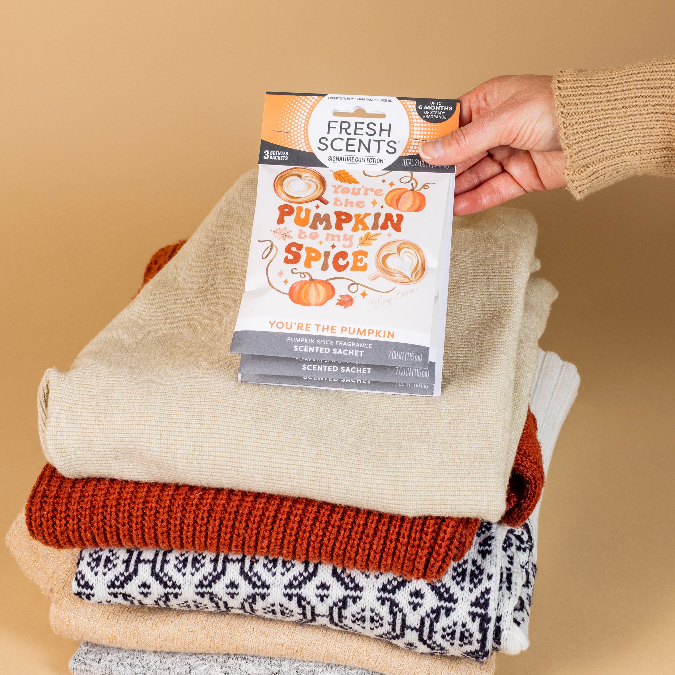 A hand places the Fresh Scents You're the Pumpkin Sachet 3pk on folded neutral sweaters, set against beige—an inviting flameless air freshener that fills your space with long-lasting pumpkin spice fragrance.