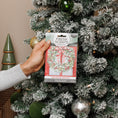 Load image into Gallery viewer, A hand holds a Fresh Scents Red Door Wreath - Sachet 3pk, a flameless fragrance, in front of a snow-frosted Christmas tree adorned with ornaments and a cone-shaped decoration nearby.
