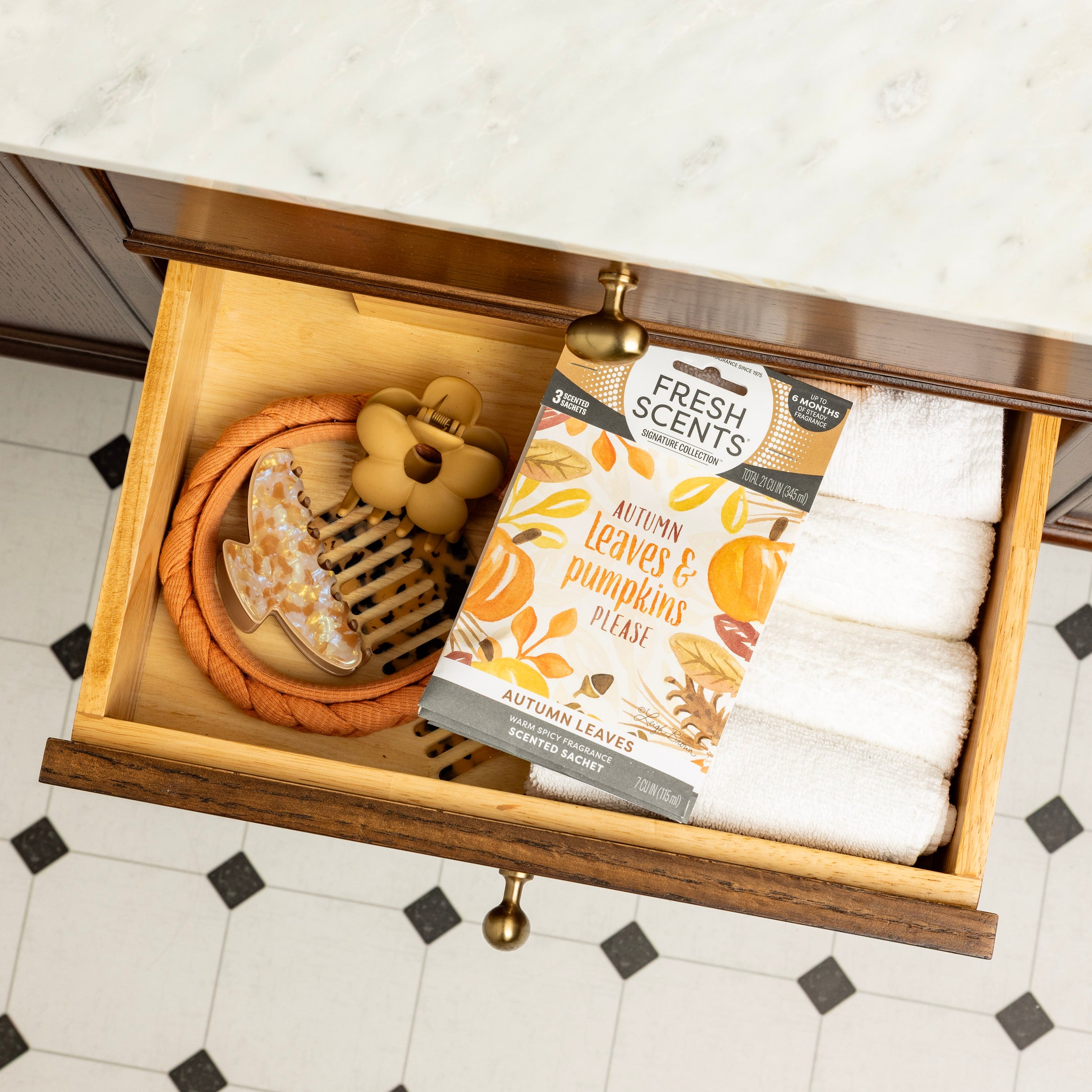 An open wooden drawer holds neatly rolled white towels, a hair clip, a wooden hairbrush, and a Fresh Scents Autumn Leaves - Sachet 3pk for long-lasting fragrance, all set on a tiled floor.