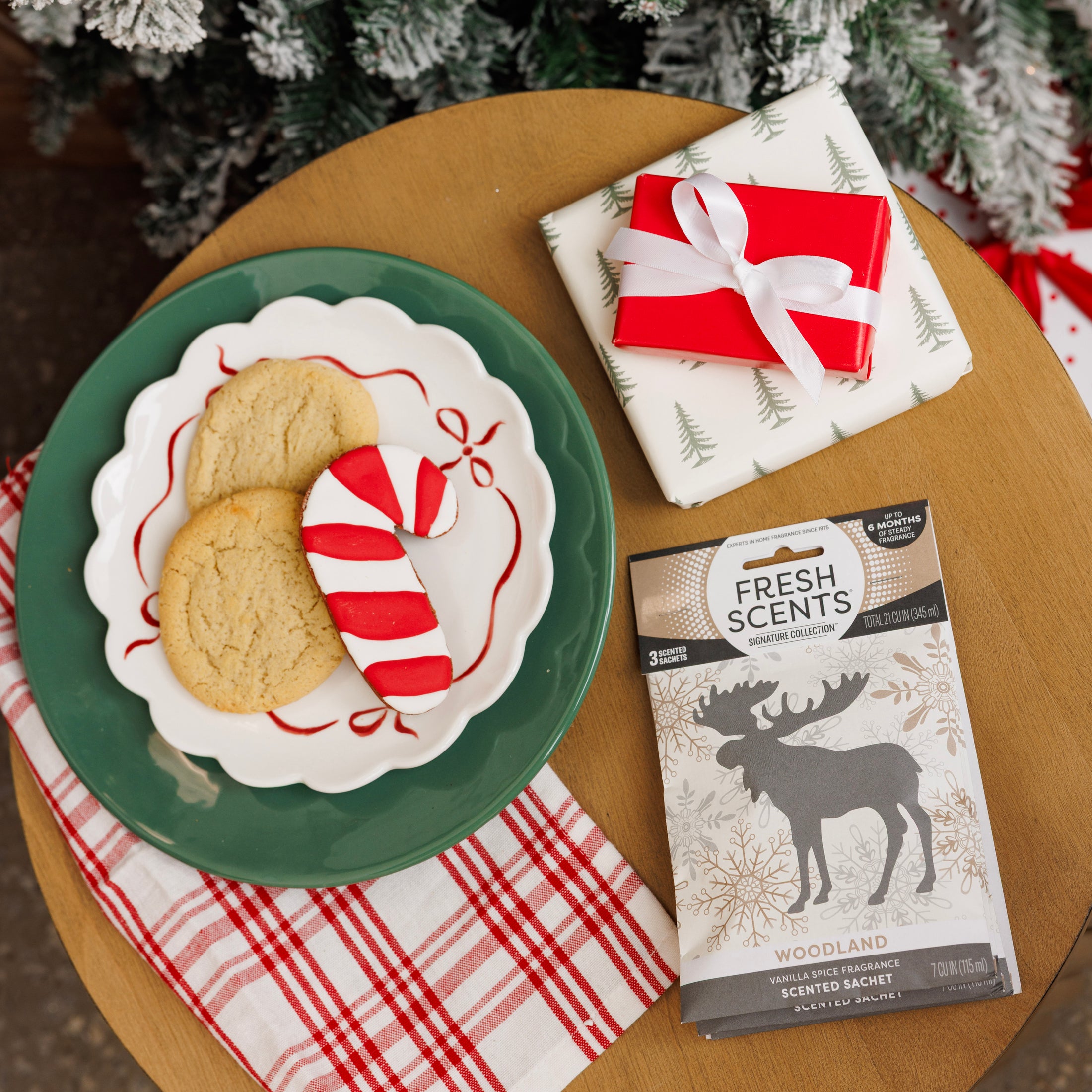 A green plate holds two sugar cookies and a candy cane cookie beside a pine tree gift-wrapped present with a red ribbon, a Fresh Scents Woodland Sachet 3pk, and a red and white napkin on a round table.