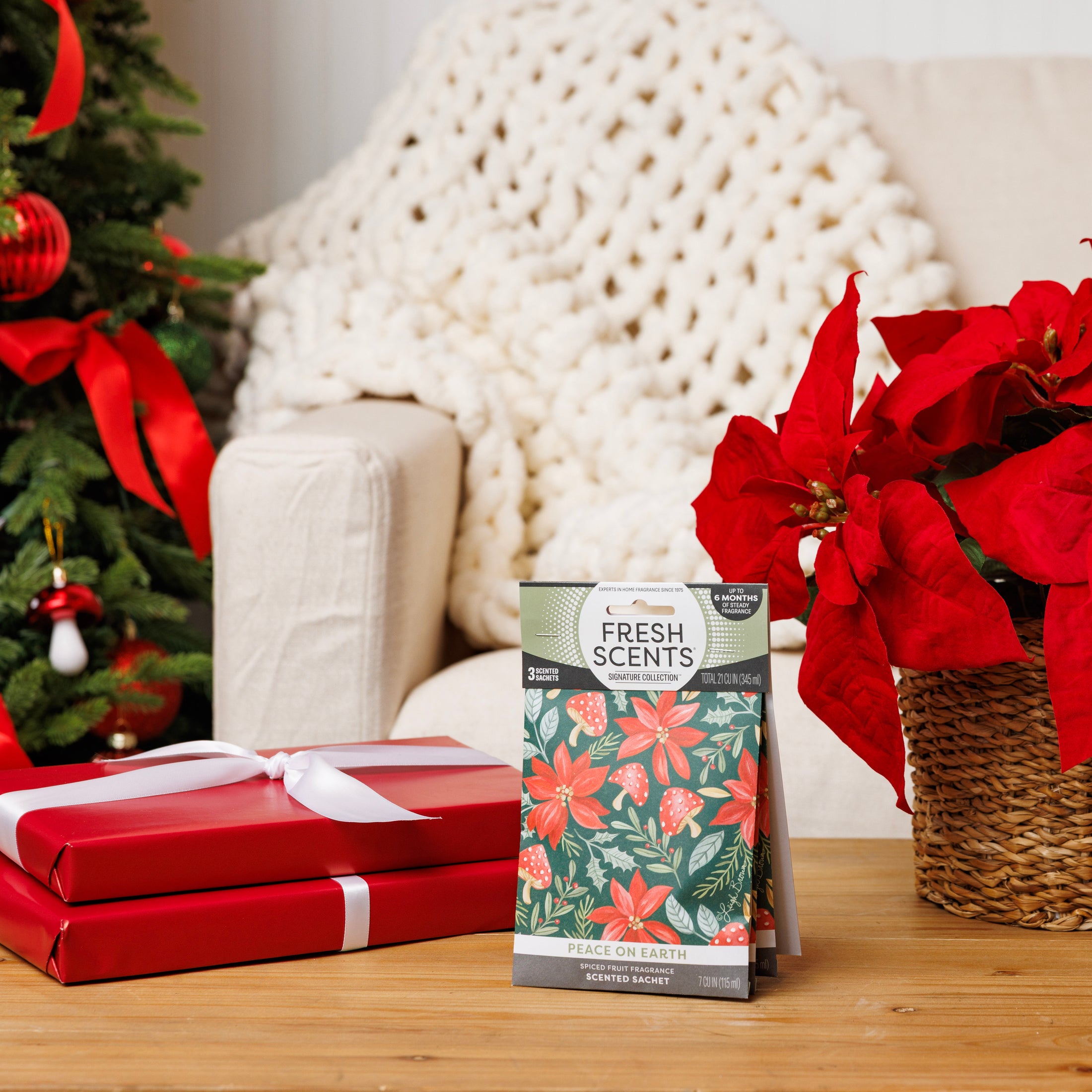 A package of Fresh Scents’ Peace on Earth - Sachet 3pk sits on a wooden table beside red poinsettias and wrapped gifts, with a Christmas tree and cream-colored knit blanket in the background. These sachets deliver long-lasting, flameless fragrance from Fresh Scents that elevates your holiday ambiance.