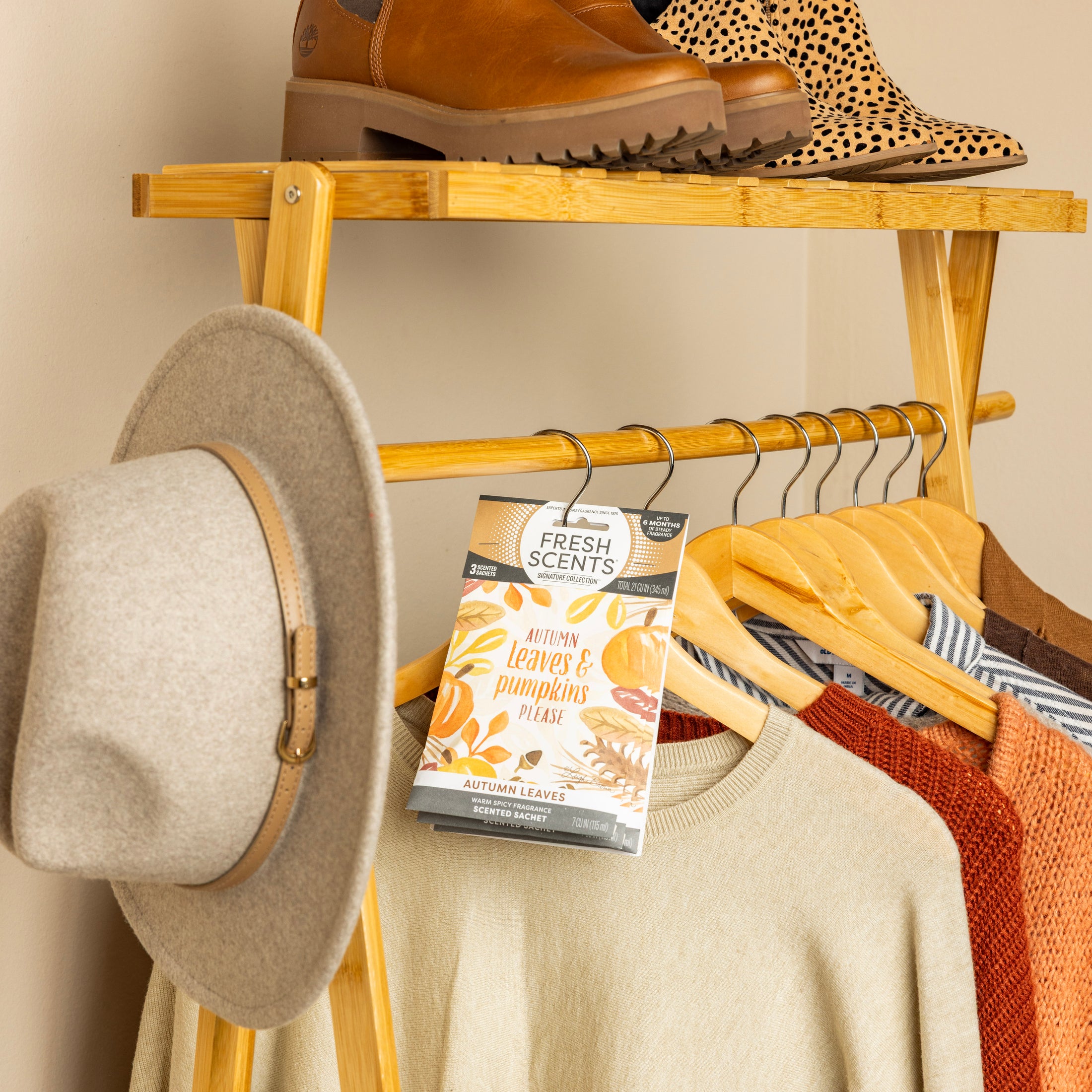A wooden clothing rack with neutral and orange sweaters, a beige hat, and shoes on the top shelf, featuring Fresh Scents Autumn Leaves - Sachet 3pk hanging on the rail for a long-lasting fall-inspired fragrance.