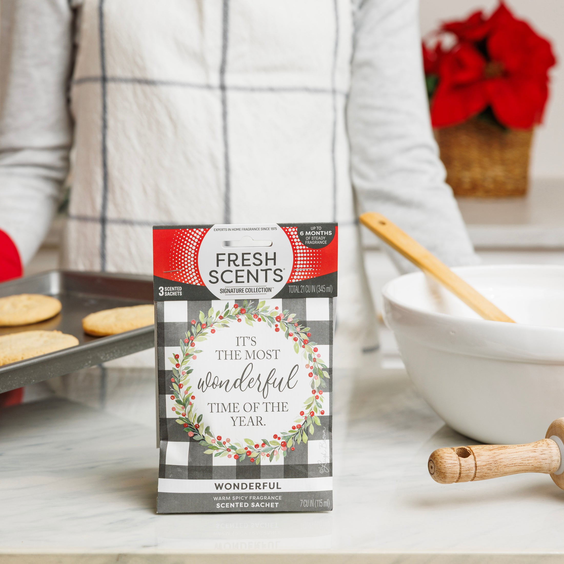 A person in an apron bakes cookies in the kitchen, while a Fresh Scents Wonderful - Sachet 3pk with a festive wreath and "It’s the most wonderful time of the year" adds a long-lasting fragrance to the scene.