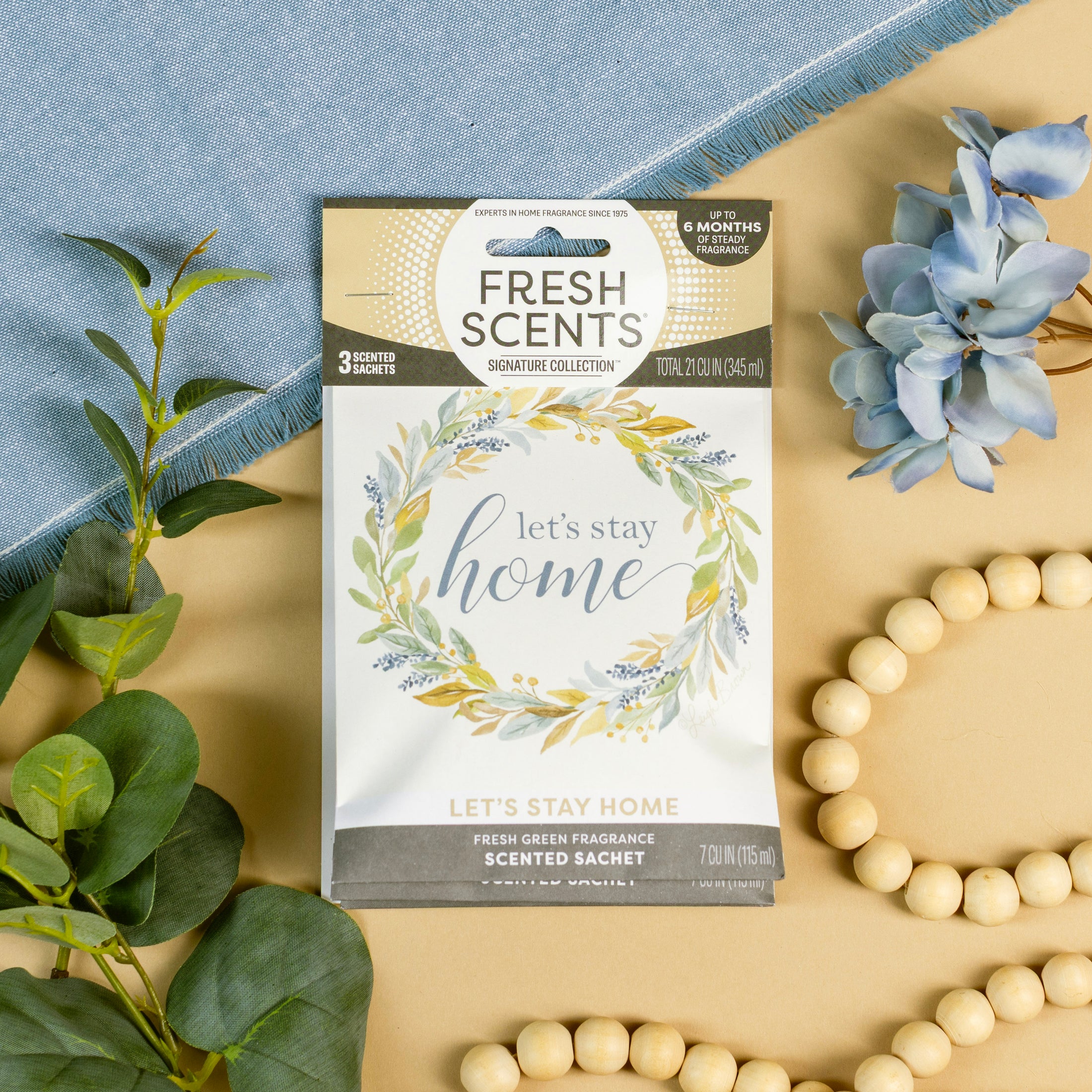 The Fresh Scents Lets Stay Home - Sachet 3pk features a floral wreath design and is displayed on beige with greenery, a beaded garland, blue cloth, and blue flowers—ideal for long-lasting fragrance from scented sachets.