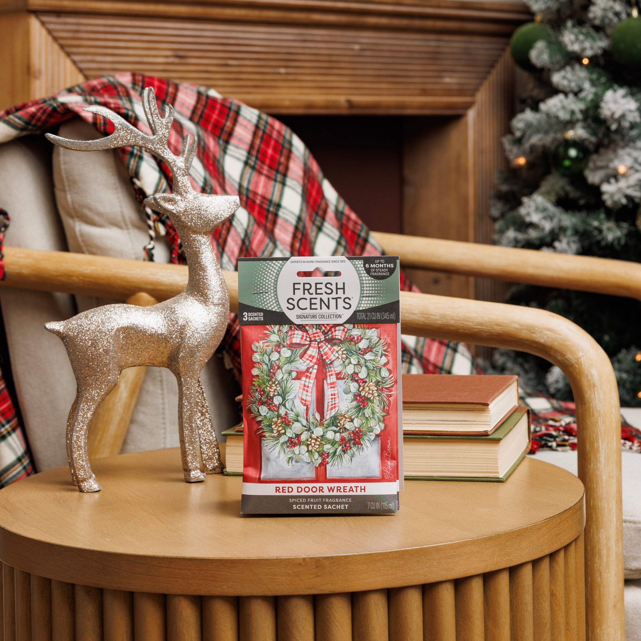 A glittery reindeer and Fresh Scents Red Door Wreath Sachet 3pk are displayed on a round wooden table with stacked books in a cozy, holiday-themed living room, featuring a plaid blanket and decorated Christmas tree.