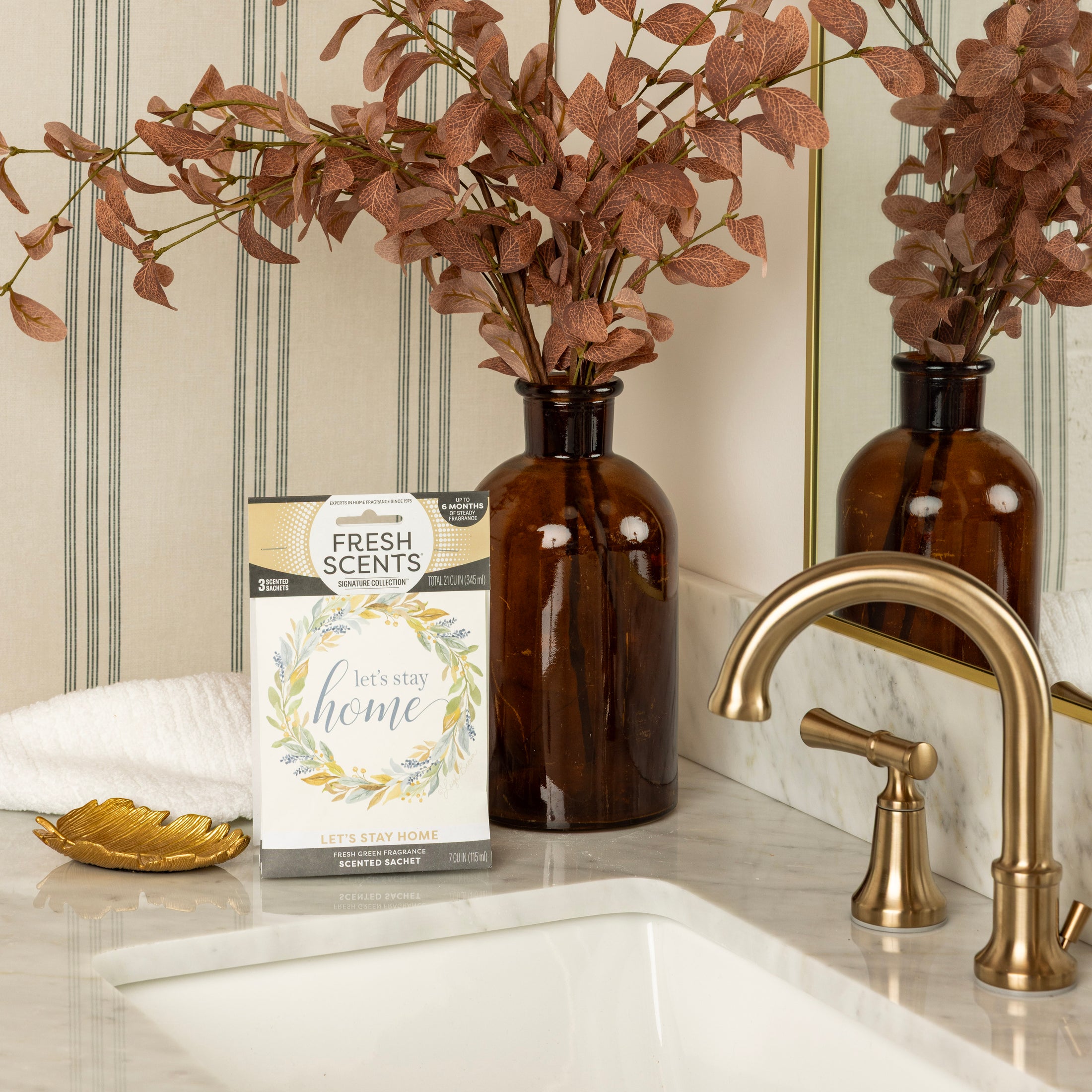 A marble bathroom counter with a brass faucet, brown glass vase with dried leaves, gold leaf-shaped dish, white towel, and Fresh Scents Lets Stay Home - Sachet 3pk for flameless fragrance and long-lasting air freshening.