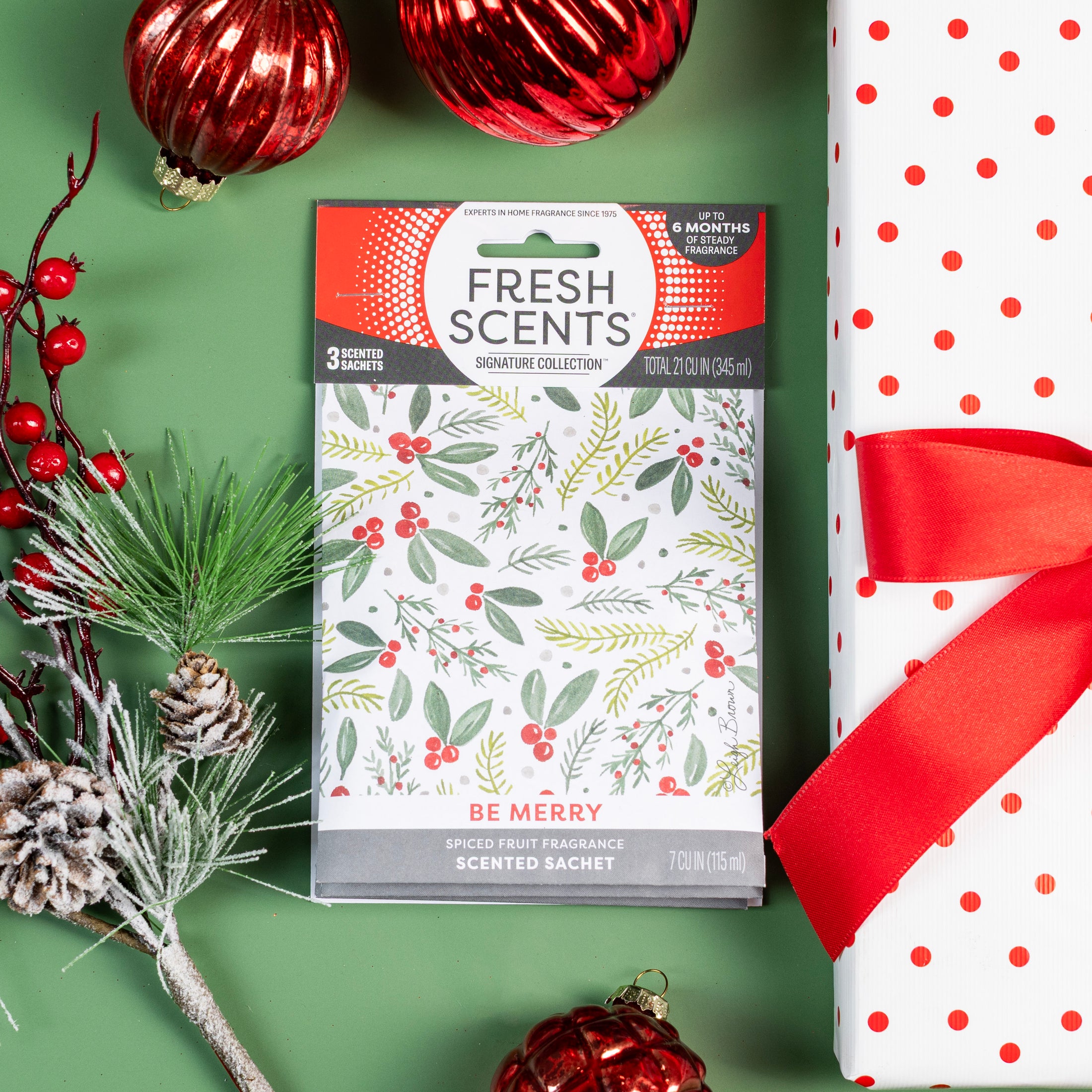 A festive scene featuring a Fresh Scents Be Merry - Sachet 3pk, red and white polka dot gift wrapped with a red ribbon, red ornaments, and a pine sprig on a green background.