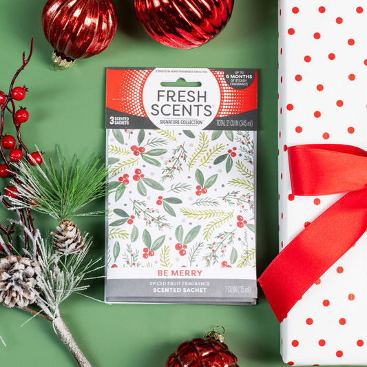 A festive scene featuring a Fresh Scents Be Merry - Sachet 3pk, red and white polka dot gift wrapped with a red ribbon, red ornaments, and a pine sprig on a green background.