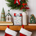 Load image into Gallery viewer, A festive mantel features ceramic houses, a Christmas tree figurine, red flowers in a vase, a wreath, and two red stockings. At the center sits Fresh Scents' Be Merry - Sachet 3pk for flameless fragrance that lasts all season.
