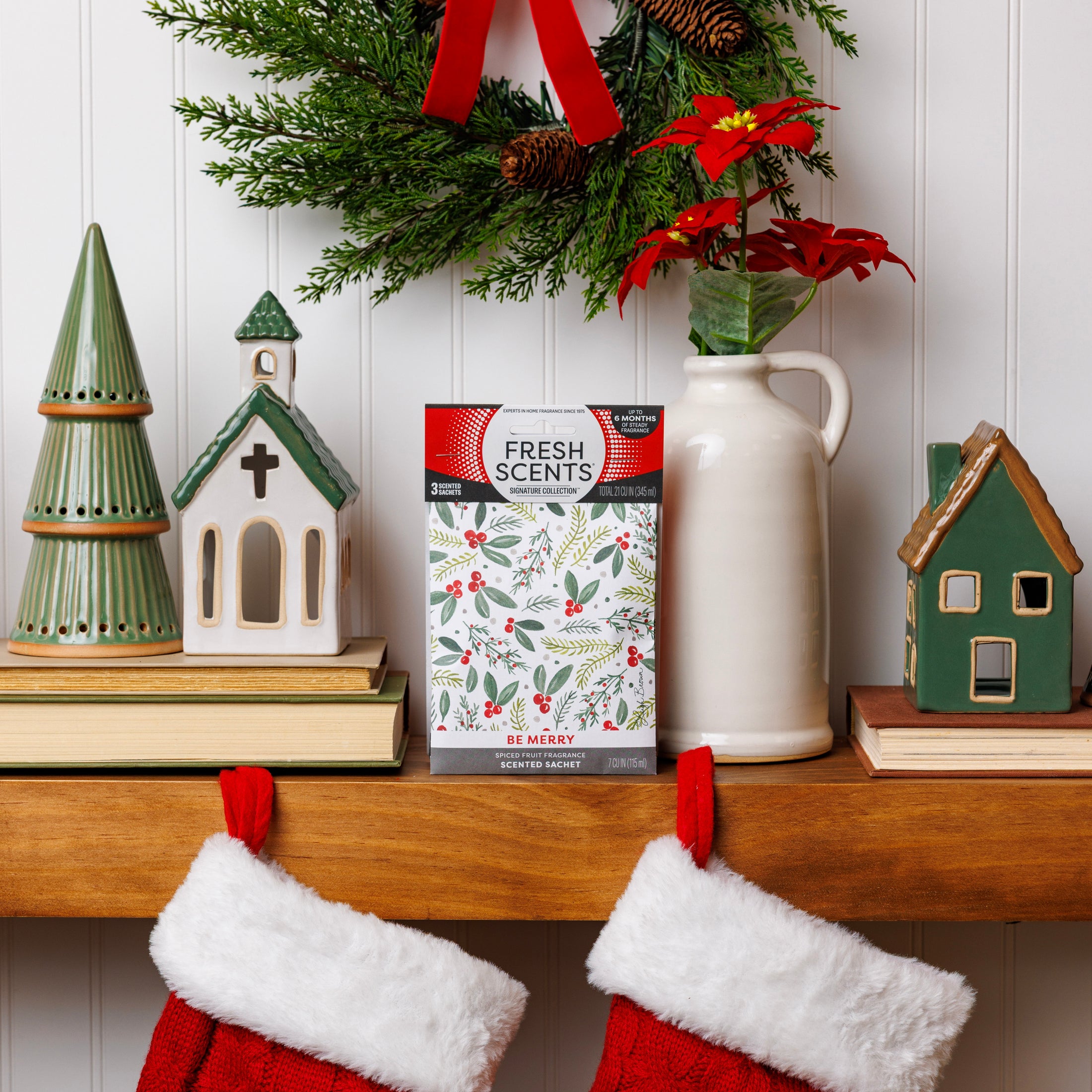 A festive mantel features ceramic houses, a Christmas tree figurine, red flowers in a vase, a wreath, and two red stockings. At the center sits Fresh Scents' Be Merry - Sachet 3pk for flameless fragrance that lasts all season.