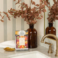 Load image into Gallery viewer, A brown glass vase with autumn leaves, a Fresh Scents Love with Fall - Sachet 3pk, a white towel, and a gold leaf dish sit on a bathroom counter with a gold faucet and striped wallpaper.
