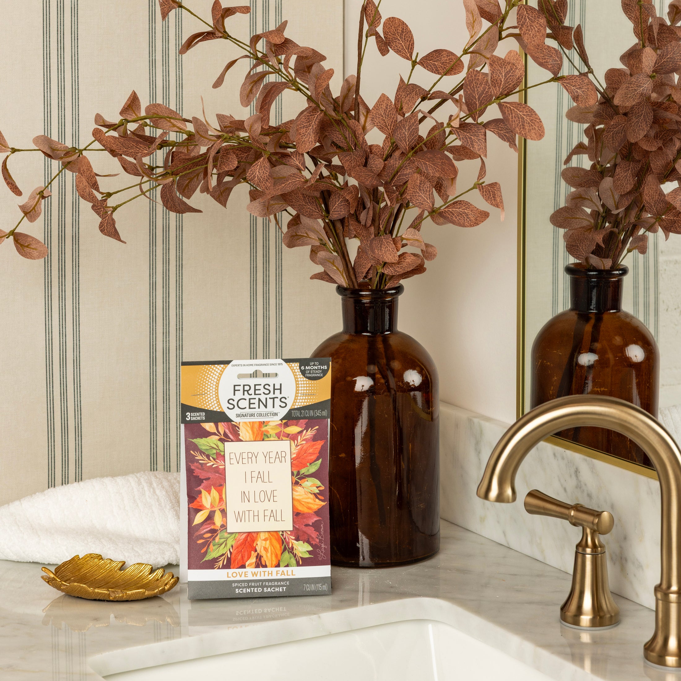 A brown glass vase with autumn leaves, a Fresh Scents Love with Fall - Sachet 3pk, a white towel, and a gold leaf dish sit on a bathroom counter with a gold faucet and striped wallpaper.