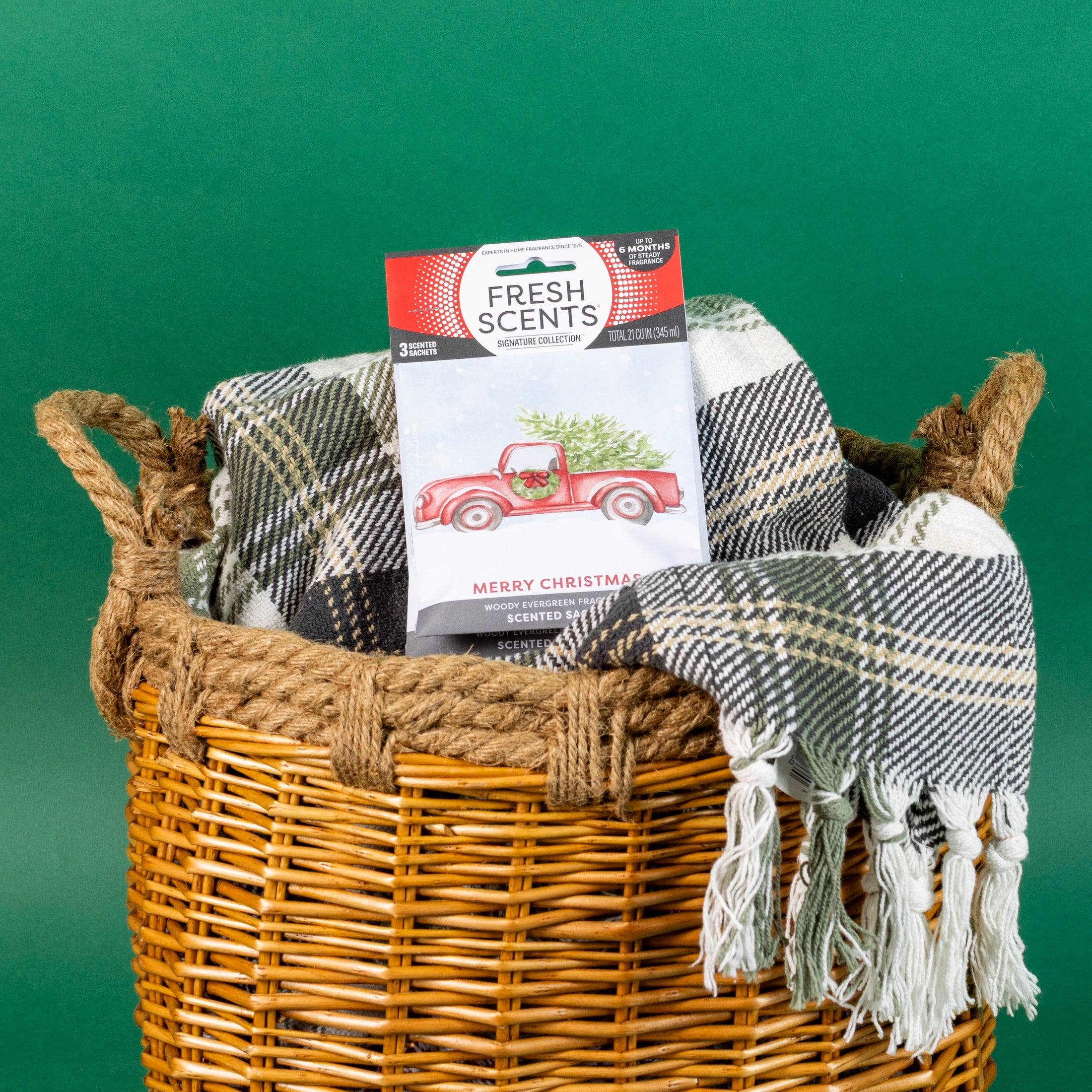 A woven basket holds a plaid fringed blanket and a Fresh Scents Merry Christmas - Sachet 3pk featuring a red truck and tree design, providing long-lasting flameless fragrance, set against a green background.