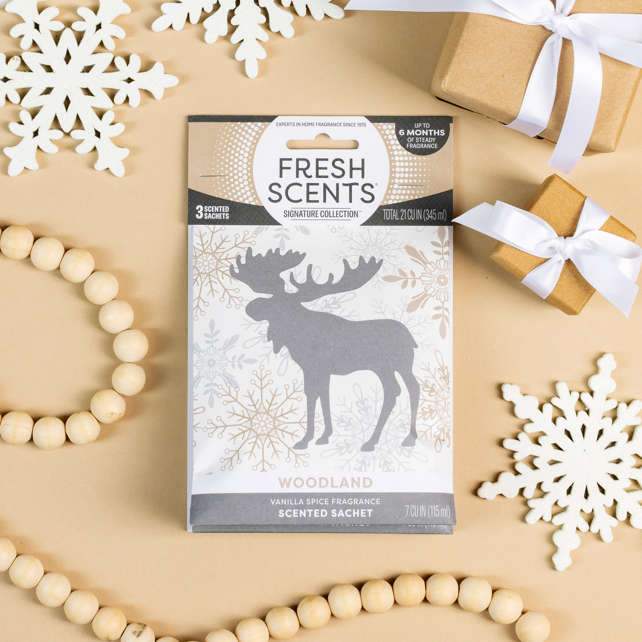 The Fresh Scents Woodland Sachet 3pk delivers a flameless vanilla spice fragrance, shown with a silver moose, white paper snowflakes, ribbon-wrapped gifts, and a wooden bead garland on a beige background.