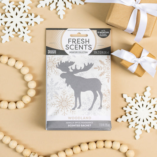 The Fresh Scents Woodland Sachet 3pk delivers a flameless vanilla spice fragrance, shown with a silver moose, white paper snowflakes, ribbon-wrapped gifts, and a wooden bead garland on a beige background.