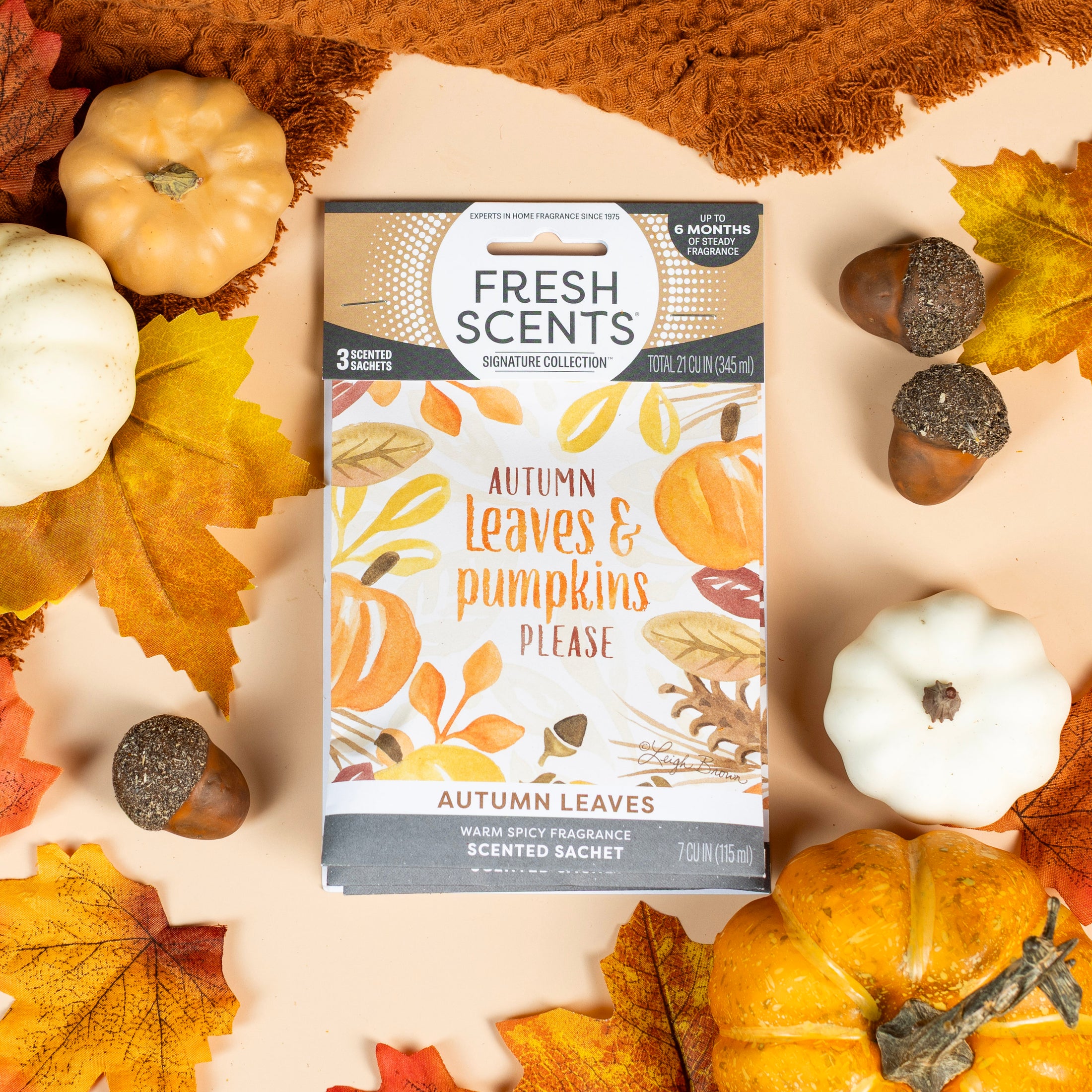 The Autumn Leaves - Sachet 3pk by Fresh Scents rests on a beige surface among autumn leaves, pumpkins, and acorns, filling the air with a cozy, long-lasting fall fragrance.