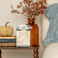 Load image into Gallery viewer, A bedside table with books, a beige knitted pumpkin, an amber vase with fall leaves, and the Fresh Scents Fall & Family - Sachet 3pk for long-lasting fragrance sits next to a bed with a blue pillow and white blanket.
