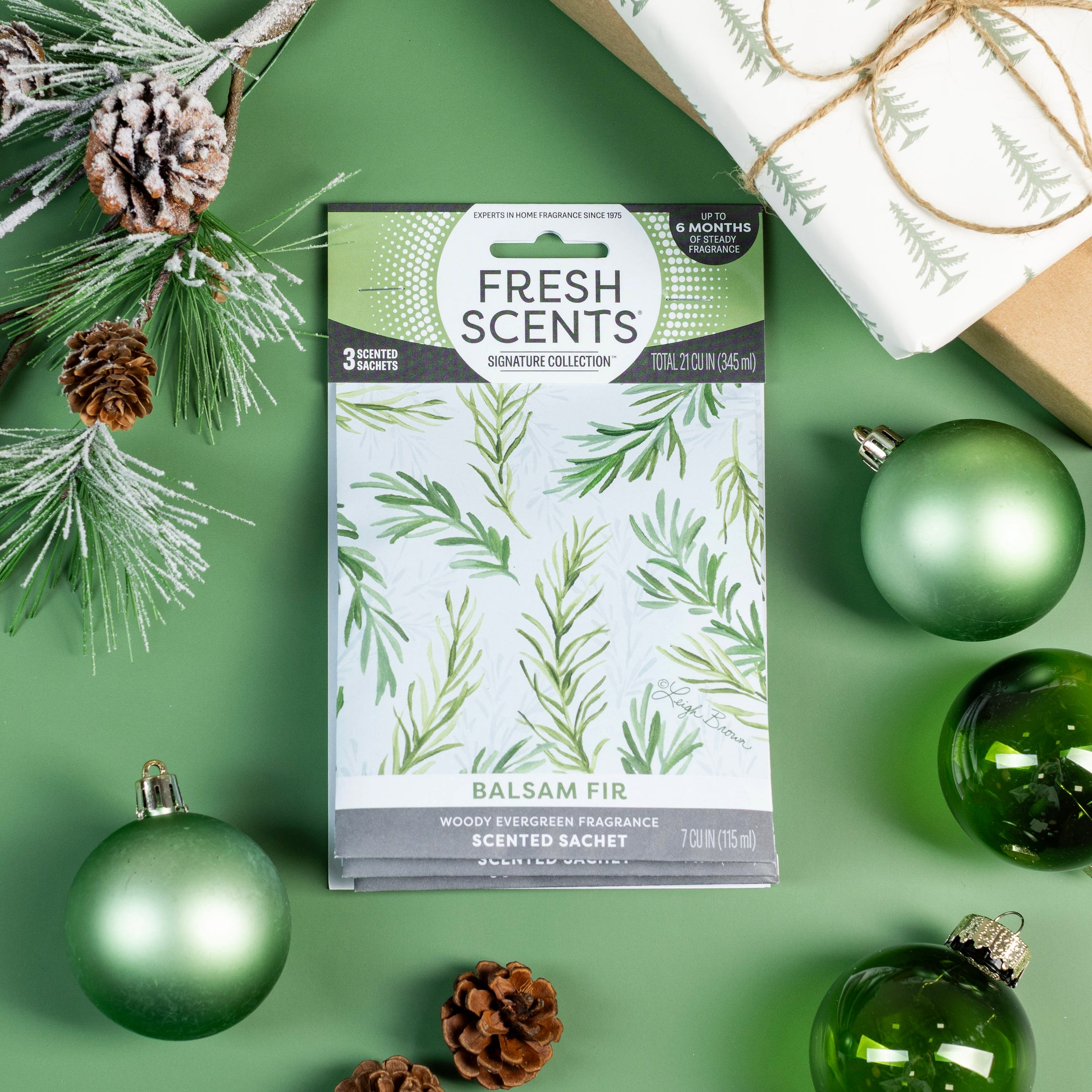A Fresh Scents Balsam Fir - Sachet 3pk rests on a green surface with pine cones, ornaments, branches, and a gift—offering long-lasting, flameless holiday fragrance to any space.