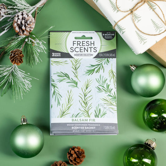 A Fresh Scents Balsam Fir - Sachet 3pk rests on a green surface with pine cones, ornaments, branches, and a gift—offering long-lasting, flameless holiday fragrance to any space.