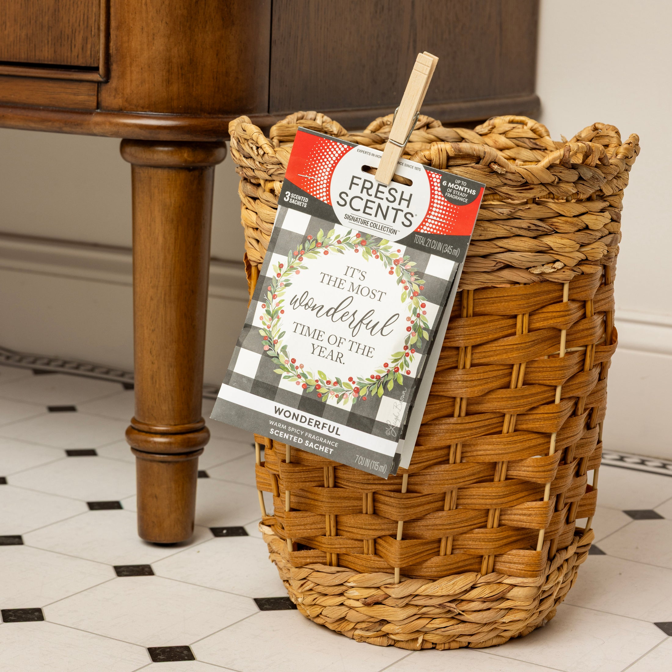 A woven wicker trash bin sits on a tiled floor by a wooden table. A Fresh Scents "Wonderful" Sachet 3pk, reading "It's the most wonderful time of the year," is clipped to the bin with a clothespin.
