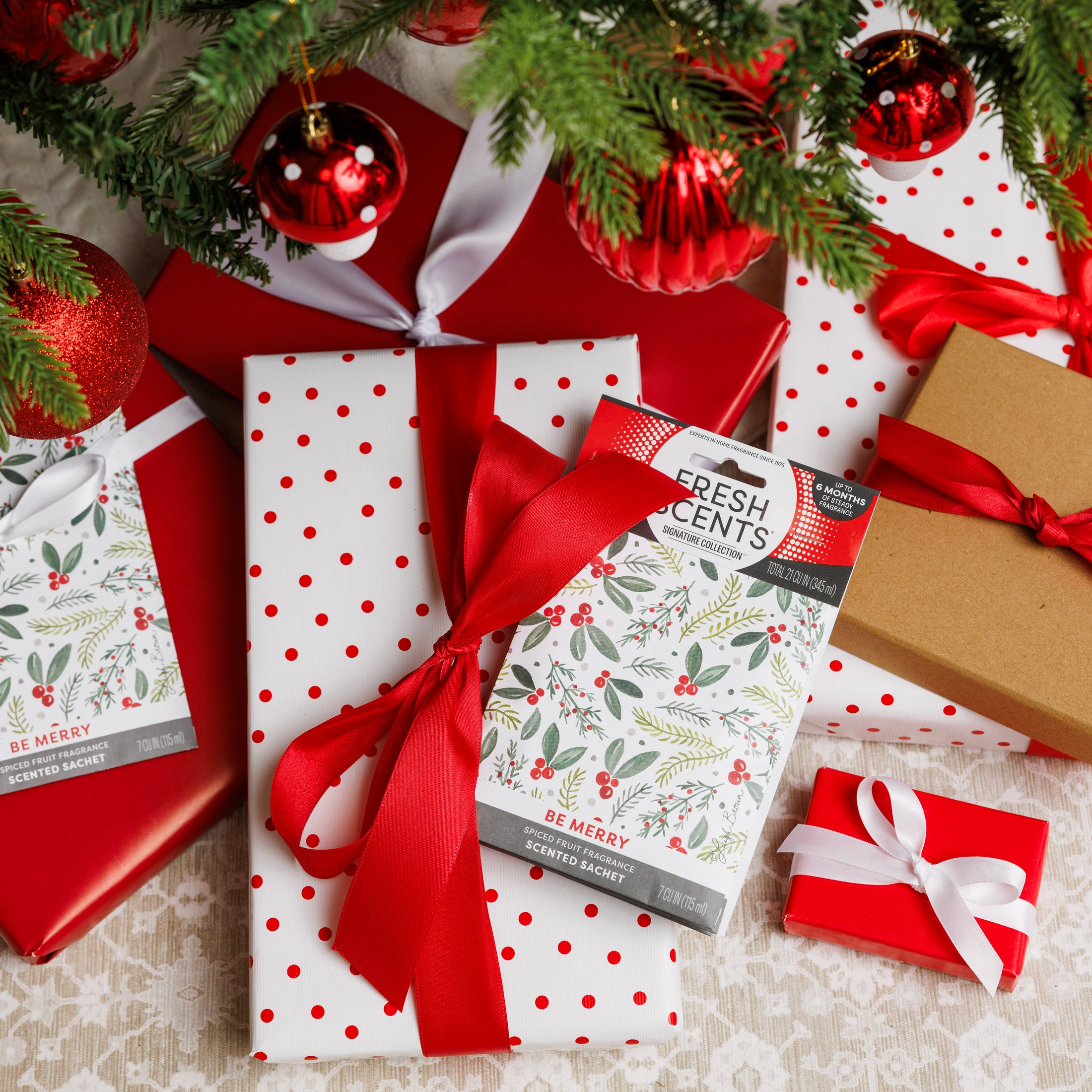 A group of Christmas presents wrapped in red, white, and brown sits under a decorated tree. One features the Fresh Scents "Be Merry" Sachet 3pk, adding a festive flameless fragrance to the cozy holiday scene.