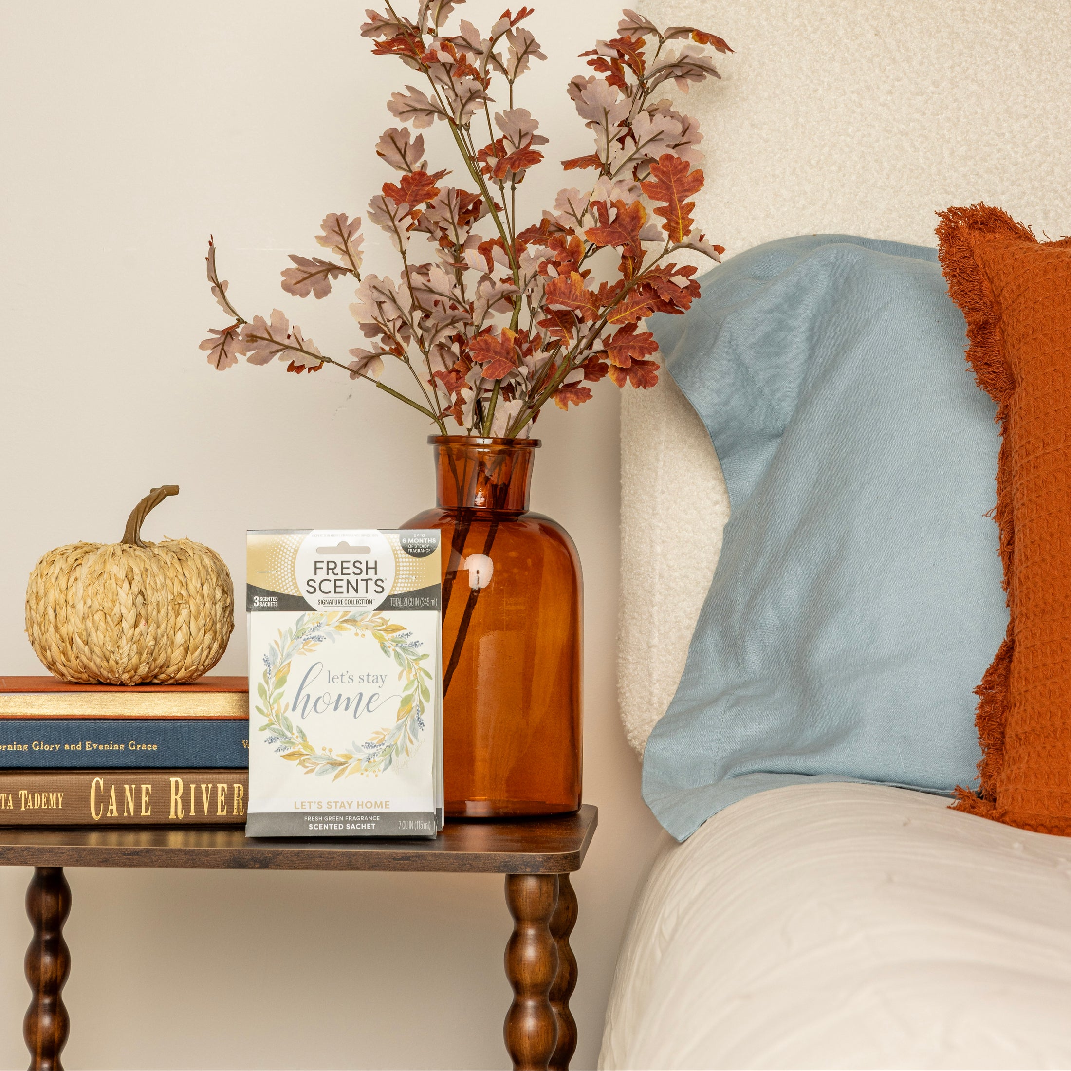 A bedside table displays a small pumpkin, two books, an amber vase with autumn leaves, and a Fresh Scents Lets Stay Home - Sachet 3pk next to a bed adorned with blue and orange pillows.