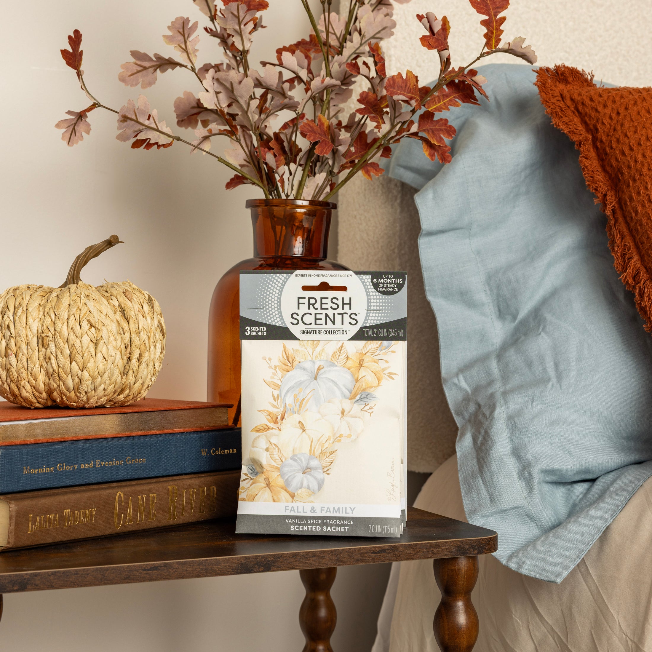 A Fresh Scents Fall & Family Sachet 3pk sits on the bedside table, surrounded by a vase of autumn leaves, a decorative pumpkin, and stacked books. A blue pillow and an orange throw blanket are nearby on the bed.