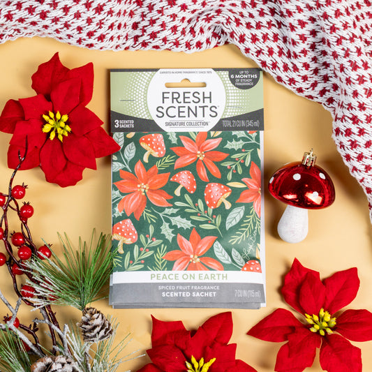 The Peace on Earth - Sachet 3pk by Fresh Scents rests on a tan surface surrounded by red poinsettias, pine branches, a cheerful red mushroom decoration, and a cozy knit red and white blanket. Featuring festive floral designs and labeled with “Peace on Earth,” these scented sachets make the perfect flameless fragrance and long-lasting freshener, beautifully infusing your space with holiday charm.