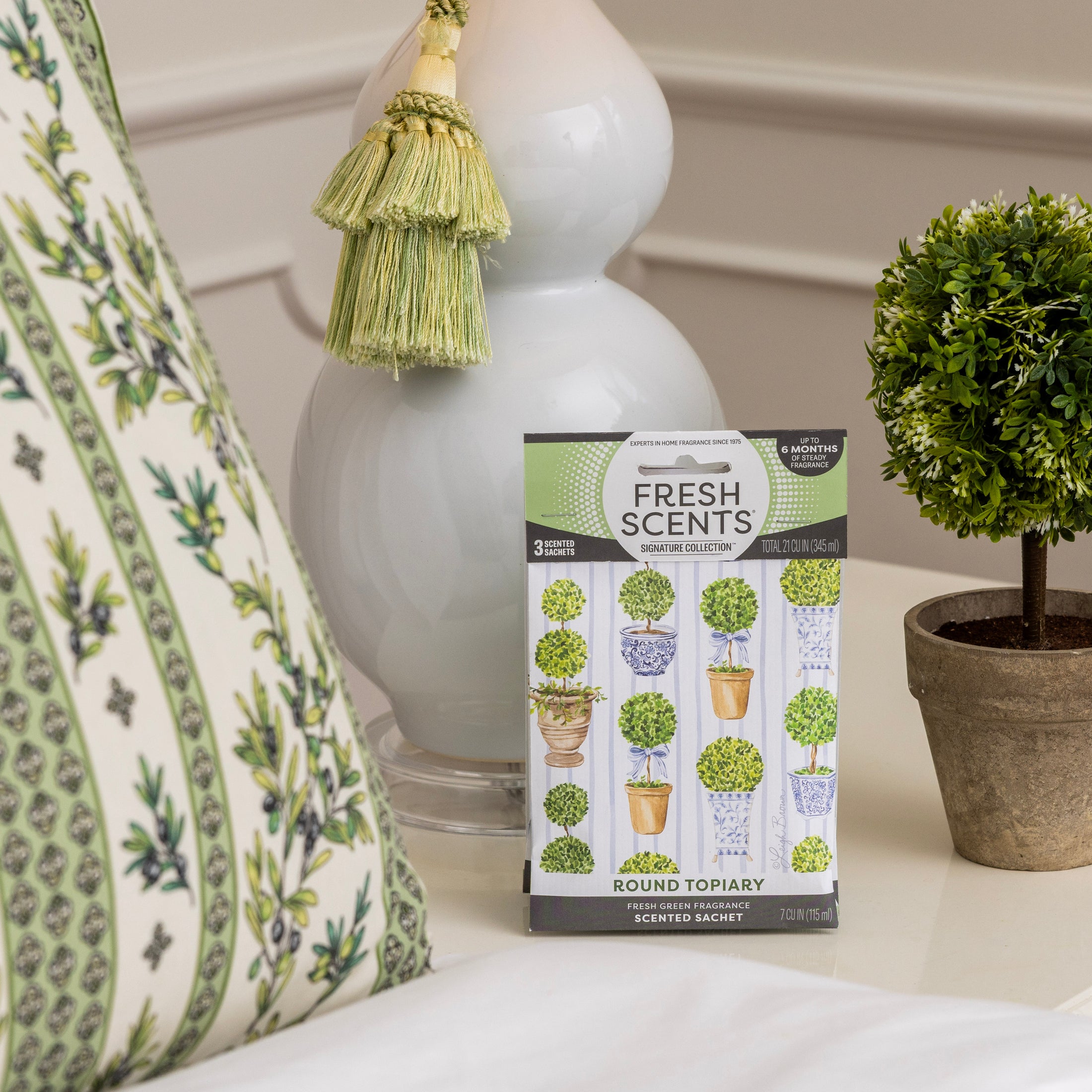 A Fresh Scents Round Topiary - Sachet 3pk, a flameless fragrance, sits on a table next to a small potted topiary and a green floral pillow. A white lamp with a green tassel appears in the background.